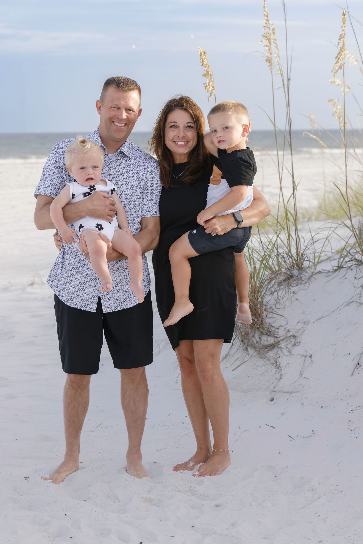 Smiling family of four on white sand beach, parents each holding a young child near sea oats.