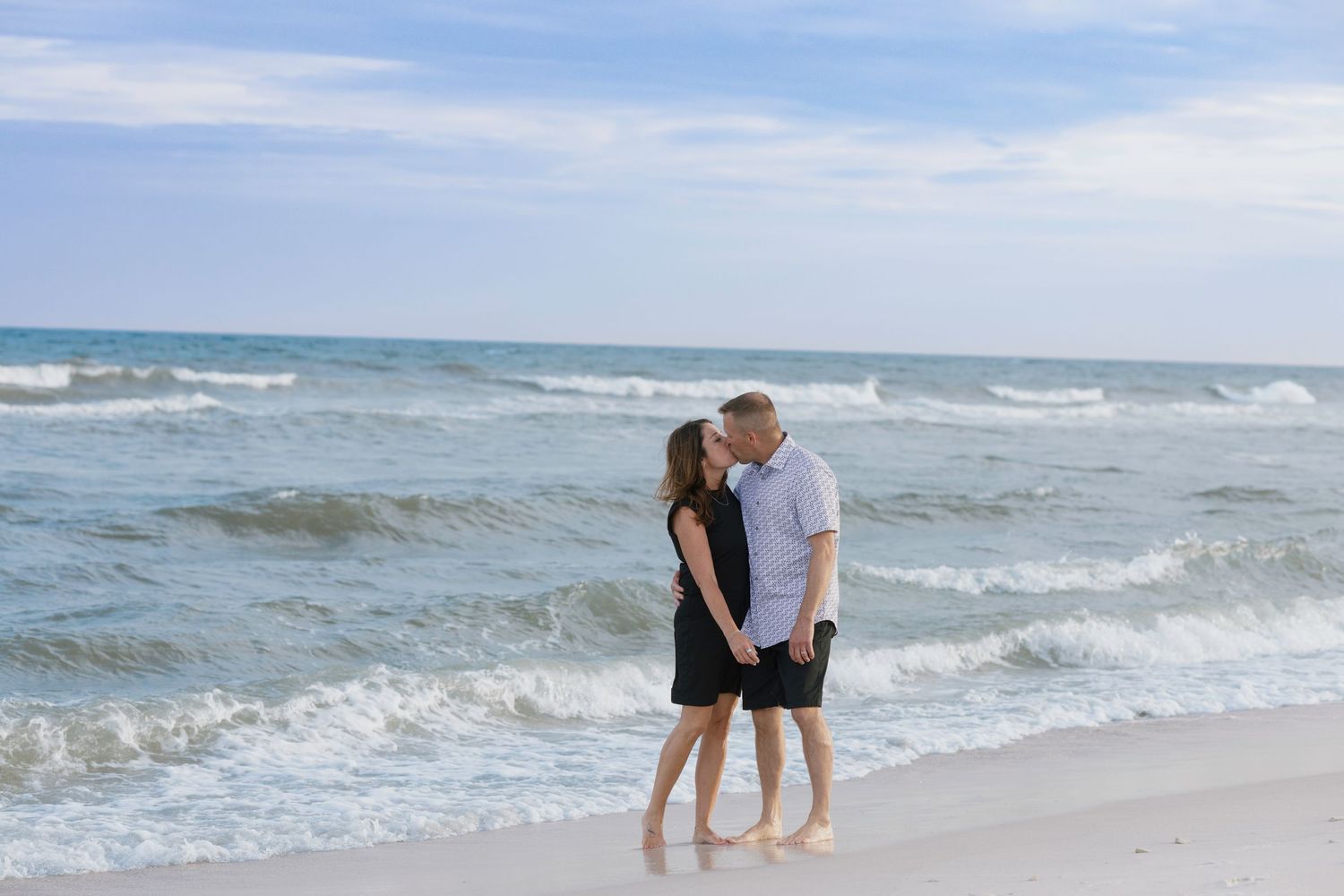 Couple sharing a romantic kiss on a sandy beach with ocean waves in the background on a clear day.