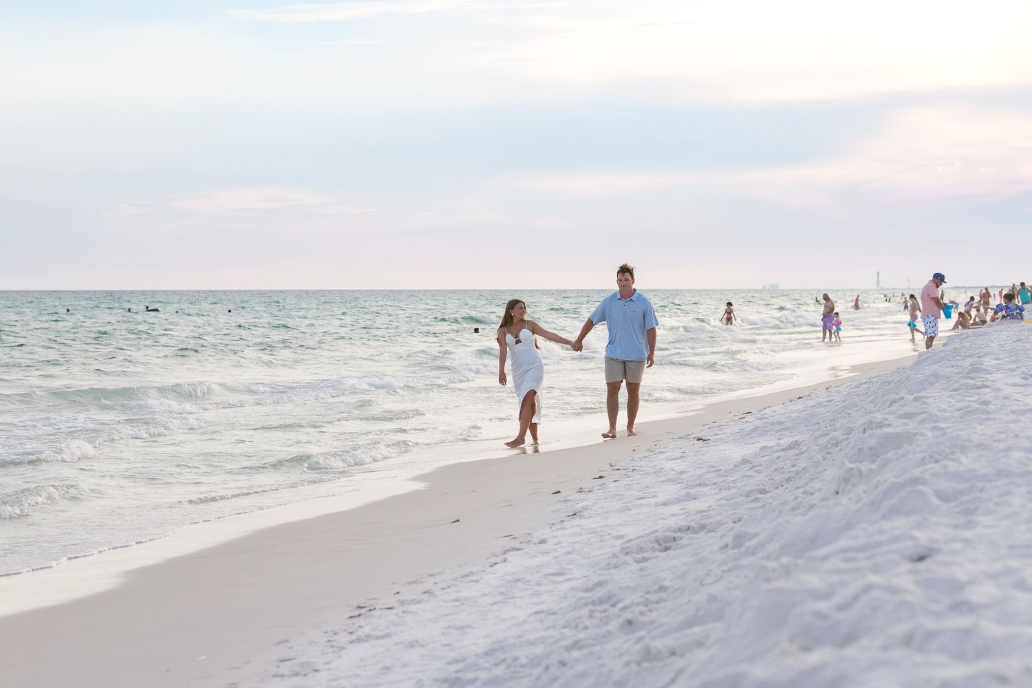 A couple holds hands walking along a white sandy beach with calm ocean waves at sunset.