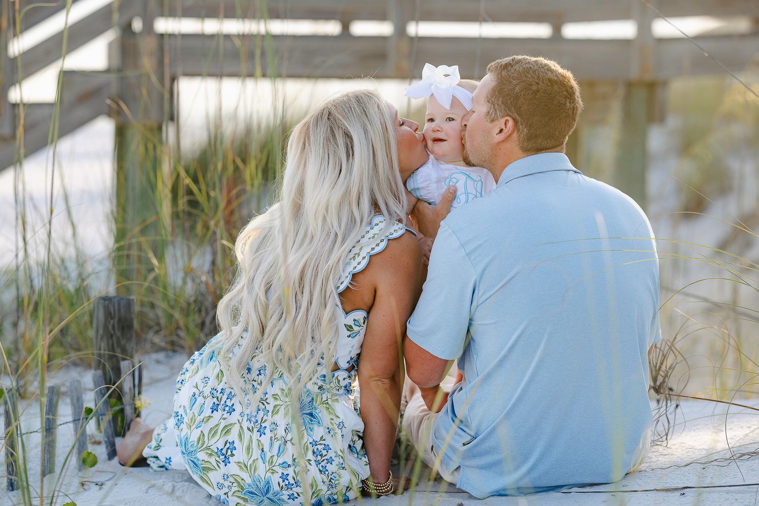 A young family sits on sandy beach steps, parents kissing their baby girl in a sweet outdoor portrait.
