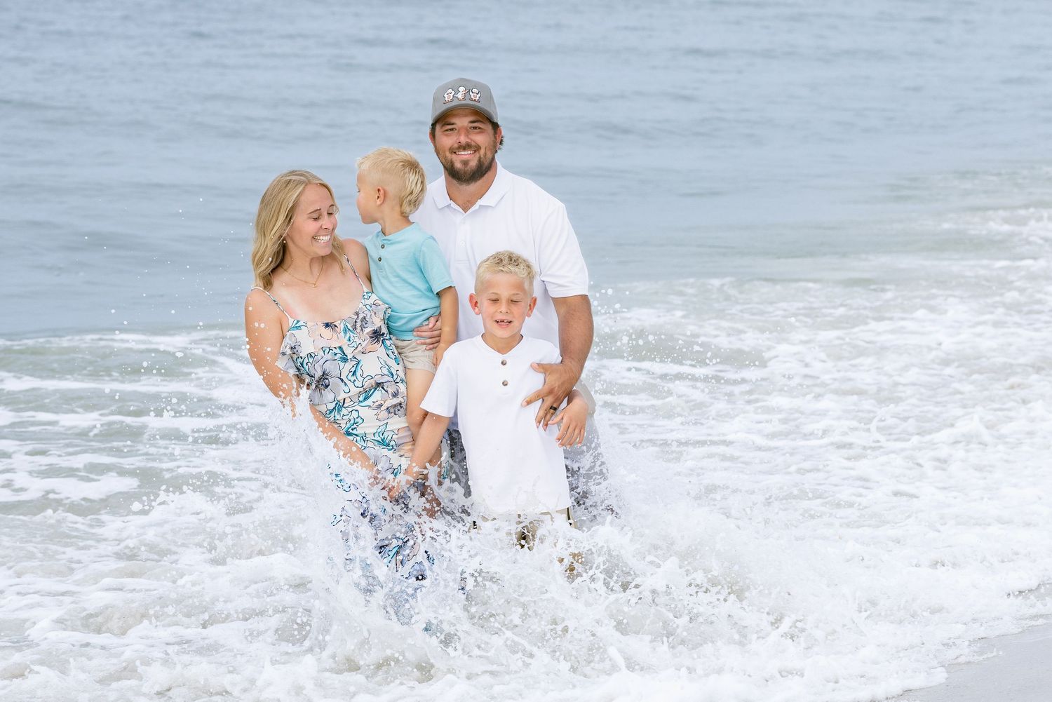 Family of four smiling and playing in ocean waves on a sunny beach day, parents holding young children.
