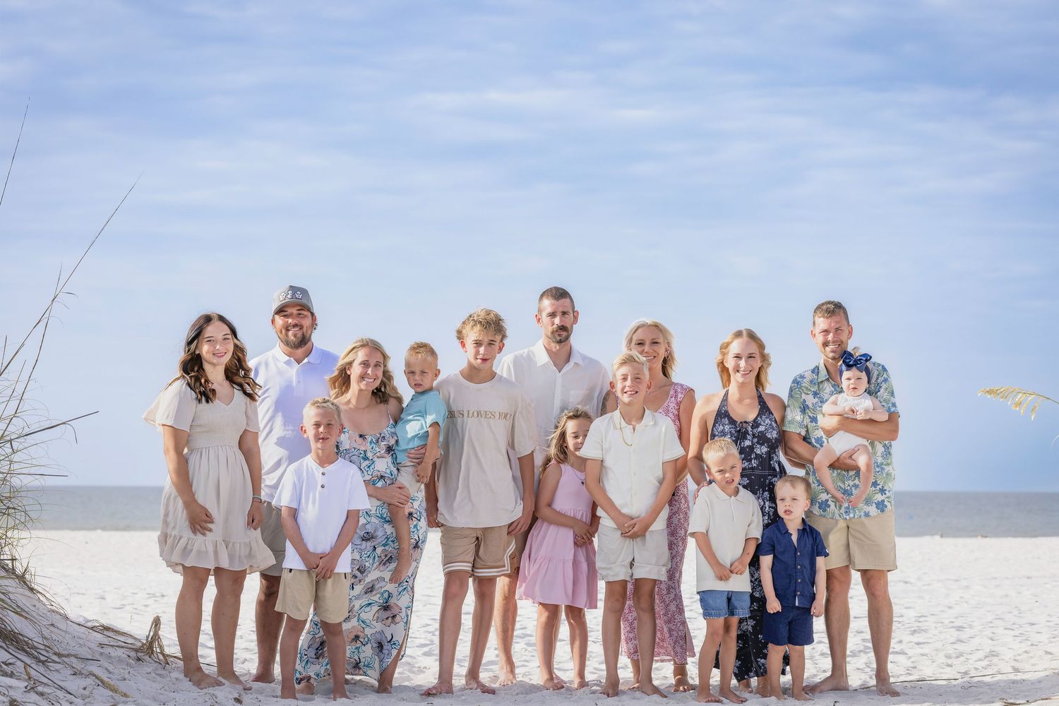 Large family group portrait on sandy beach with blue sky, adults and children smiling in coordinated pastel outfits.