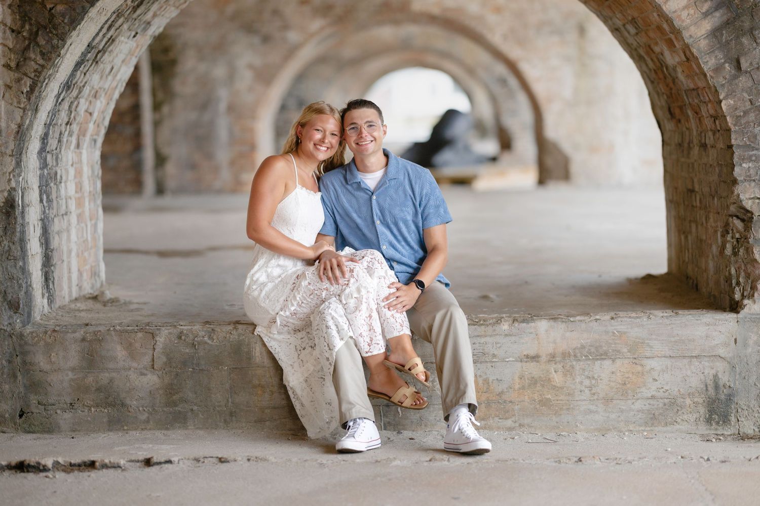 Couple sitting together under stone arches, woman in white dress and man in blue polo, smiling warmly at historic brick venue.