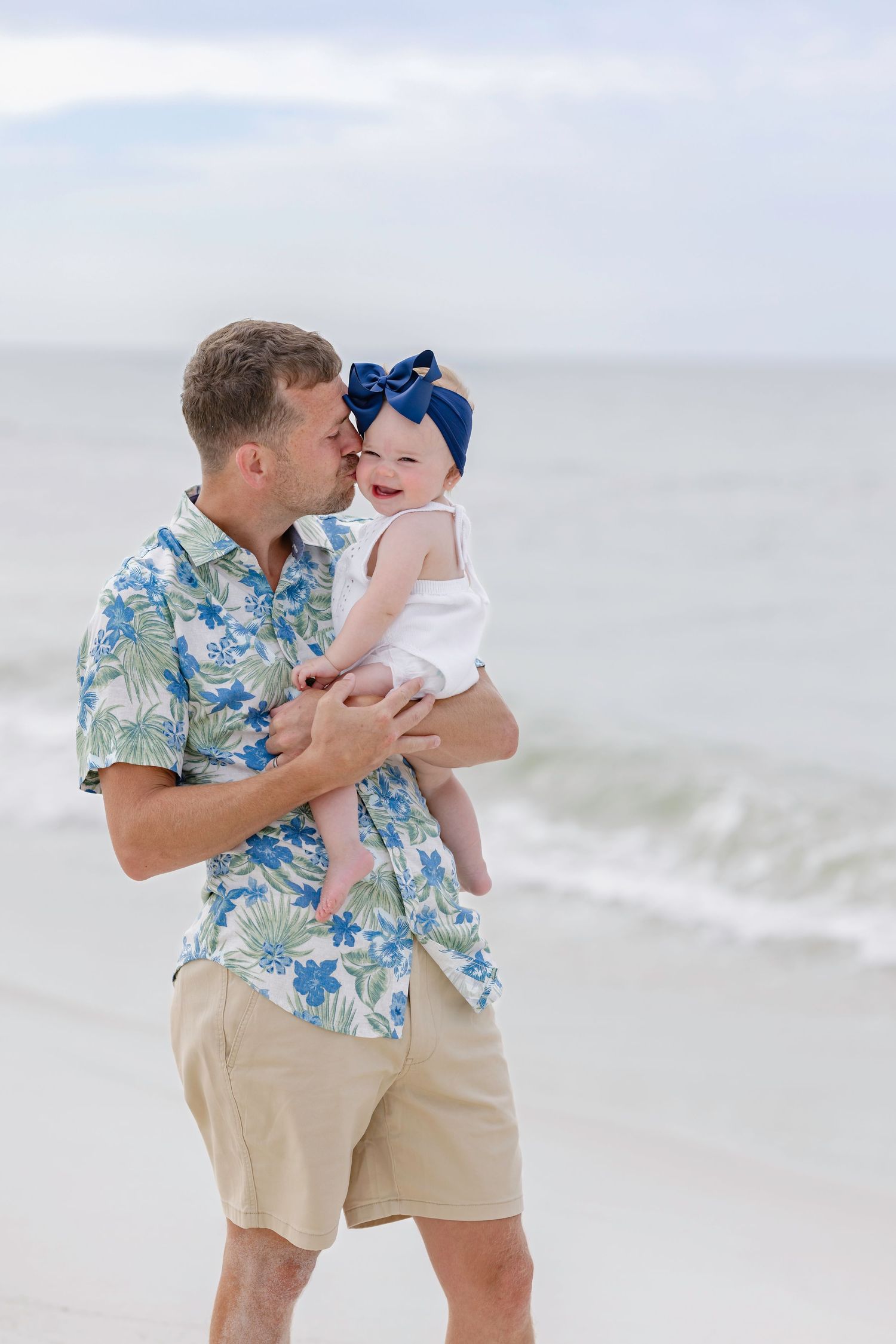 Father in floral shirt kissing his baby girl with blue bow headband on a sunny beach.