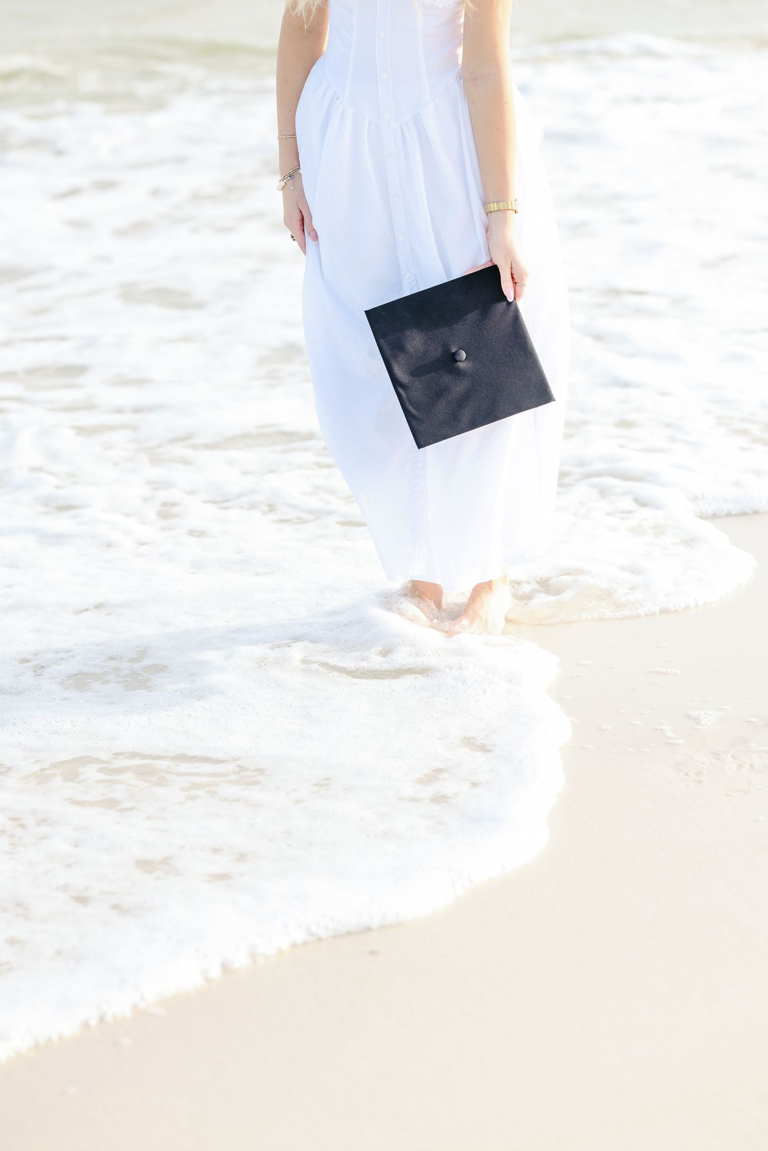 Close-up of woman in white dress standing in ocean waves holding black graduation cap.