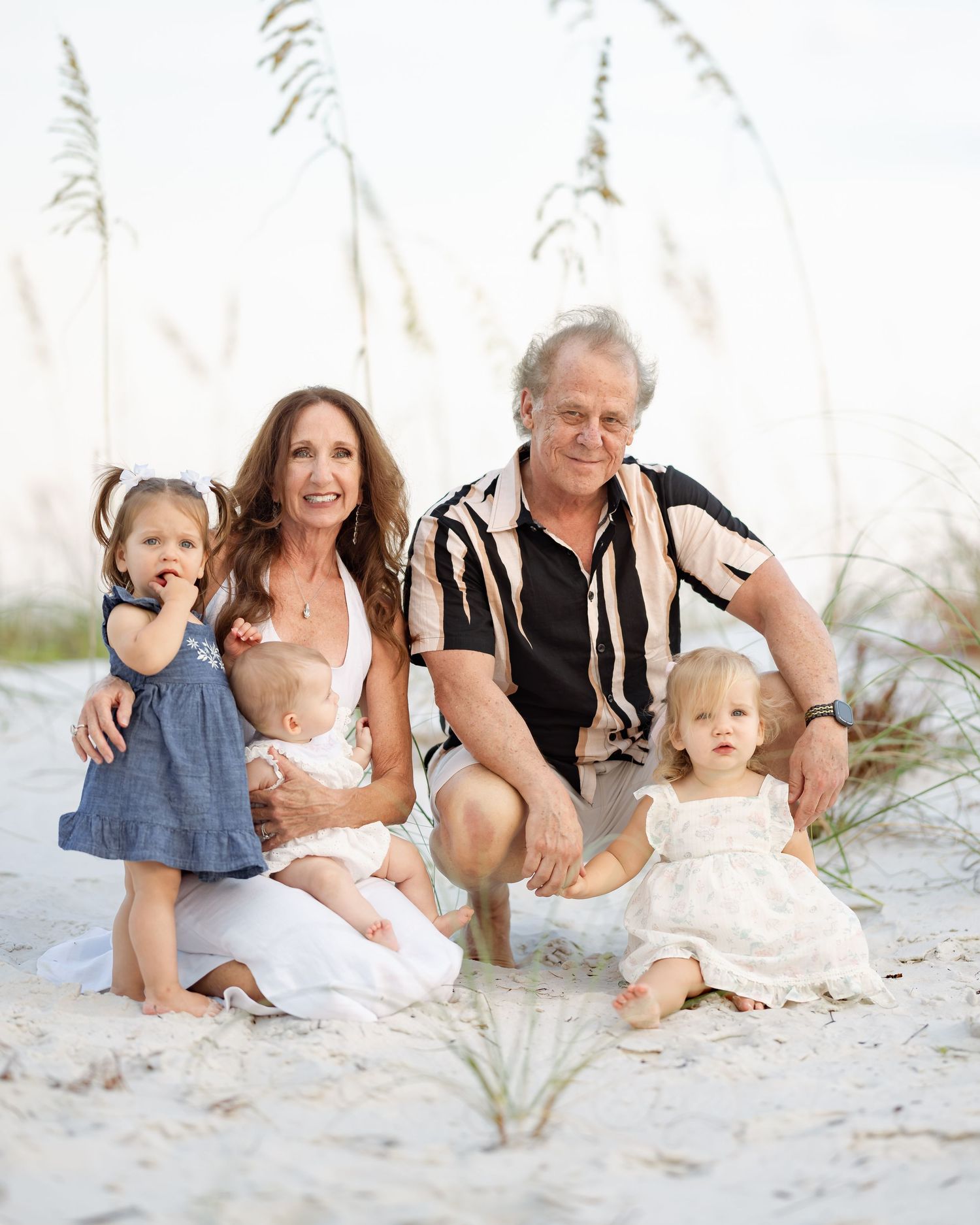 Family of five posing on sandy beach with sea oats, featuring grandparents and three young children in casual attire.