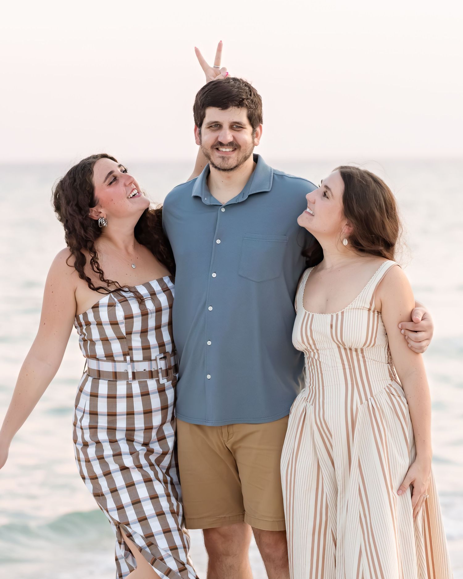 Three young adults laughing and posing together on a beach at sunset, one making bunny ears behind a smiling man.