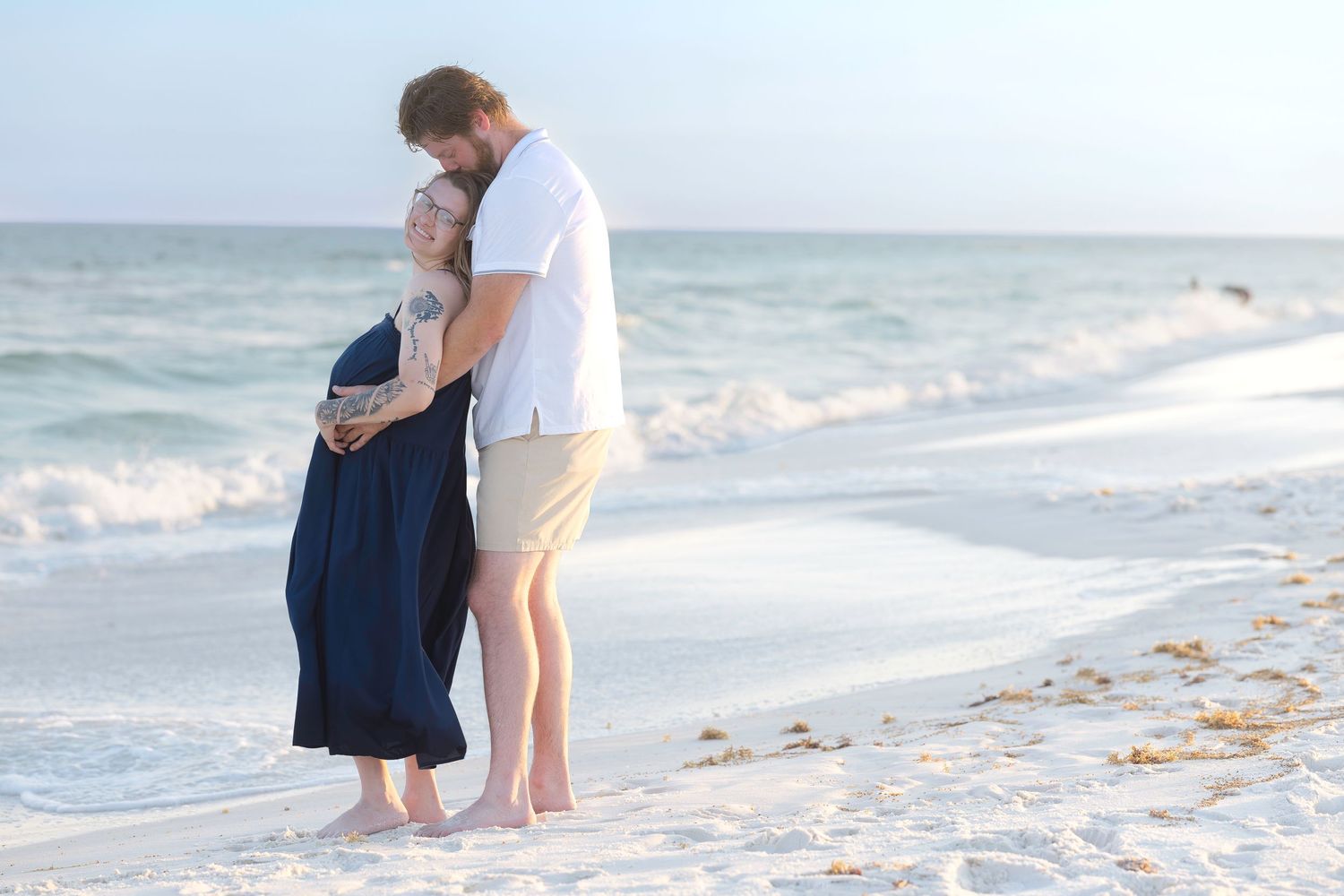 Pregnant couple embracing tenderly on a white sand beach with turquoise ocean waves.