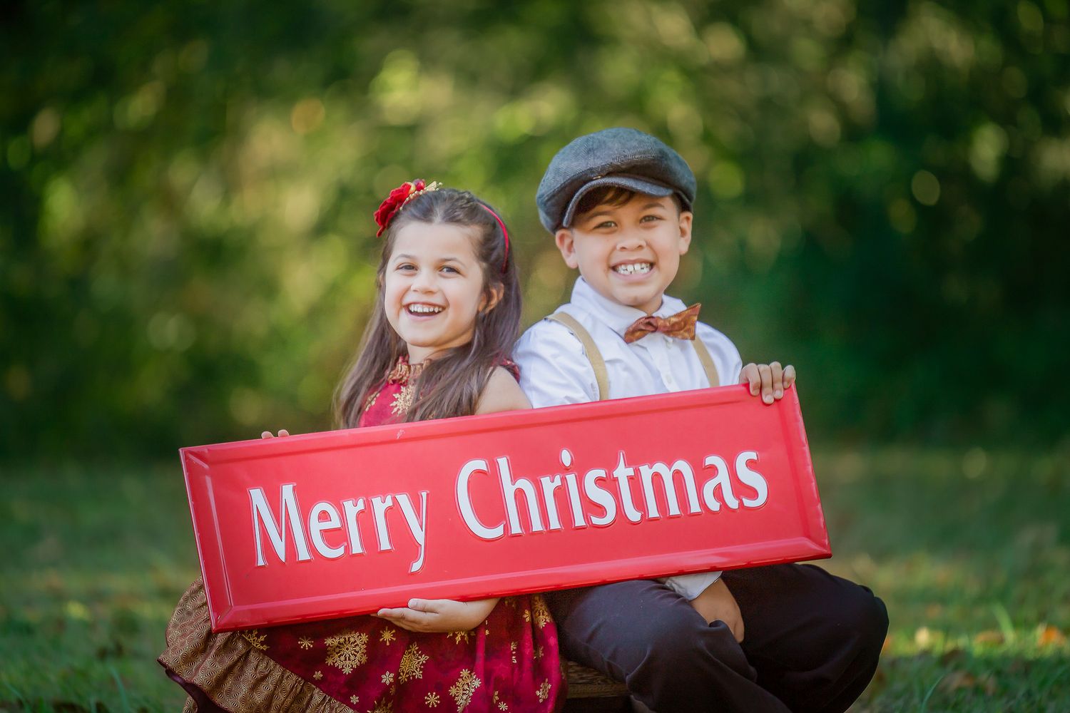 Two children in vintage-style clothing hold a red wooden Merry Christmas sign while sitting outdoors in a garden setting.
