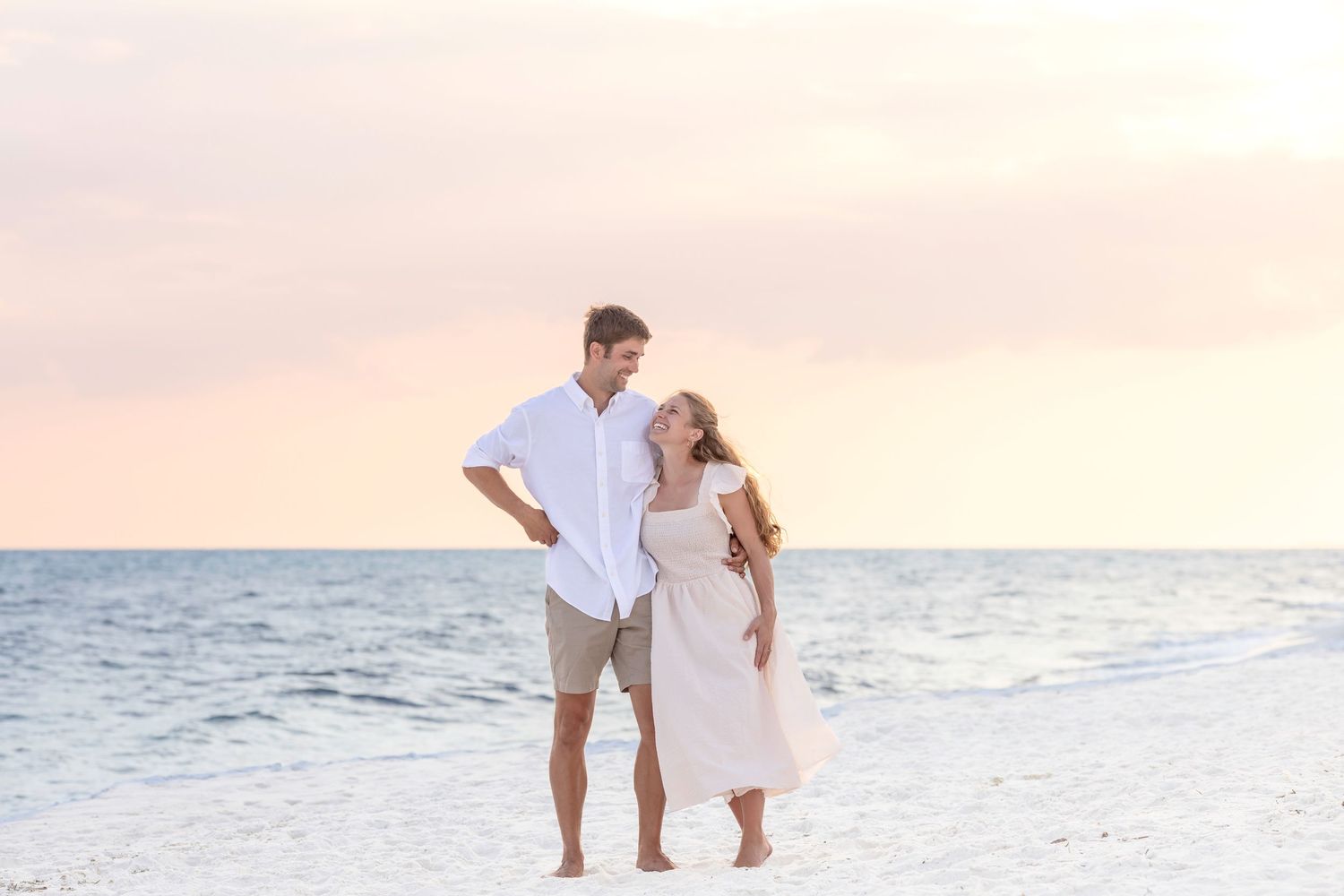 A couple in light summer clothing walks along a pristine white sandy beach at sunset.