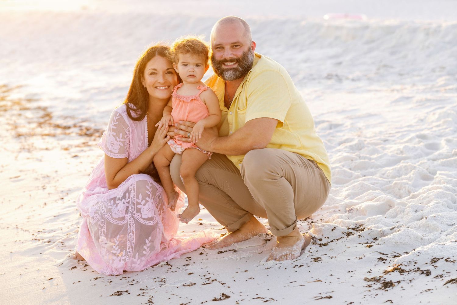 Family in pink and yellow outfits sits together on white sandy beach during golden hour sunset.