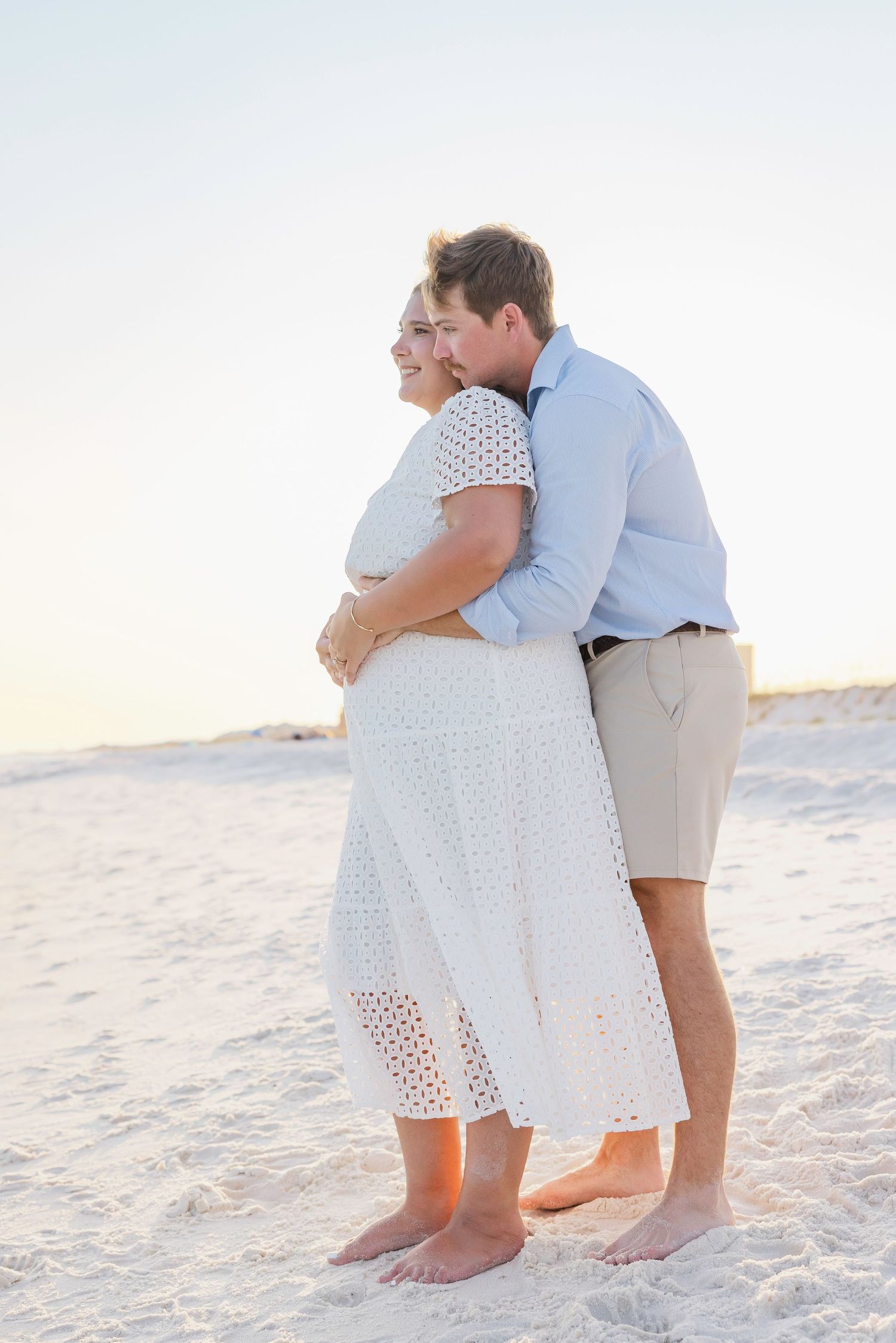 A couple embraces on a white sandy beach at sunset, wearing light summer clothing.
