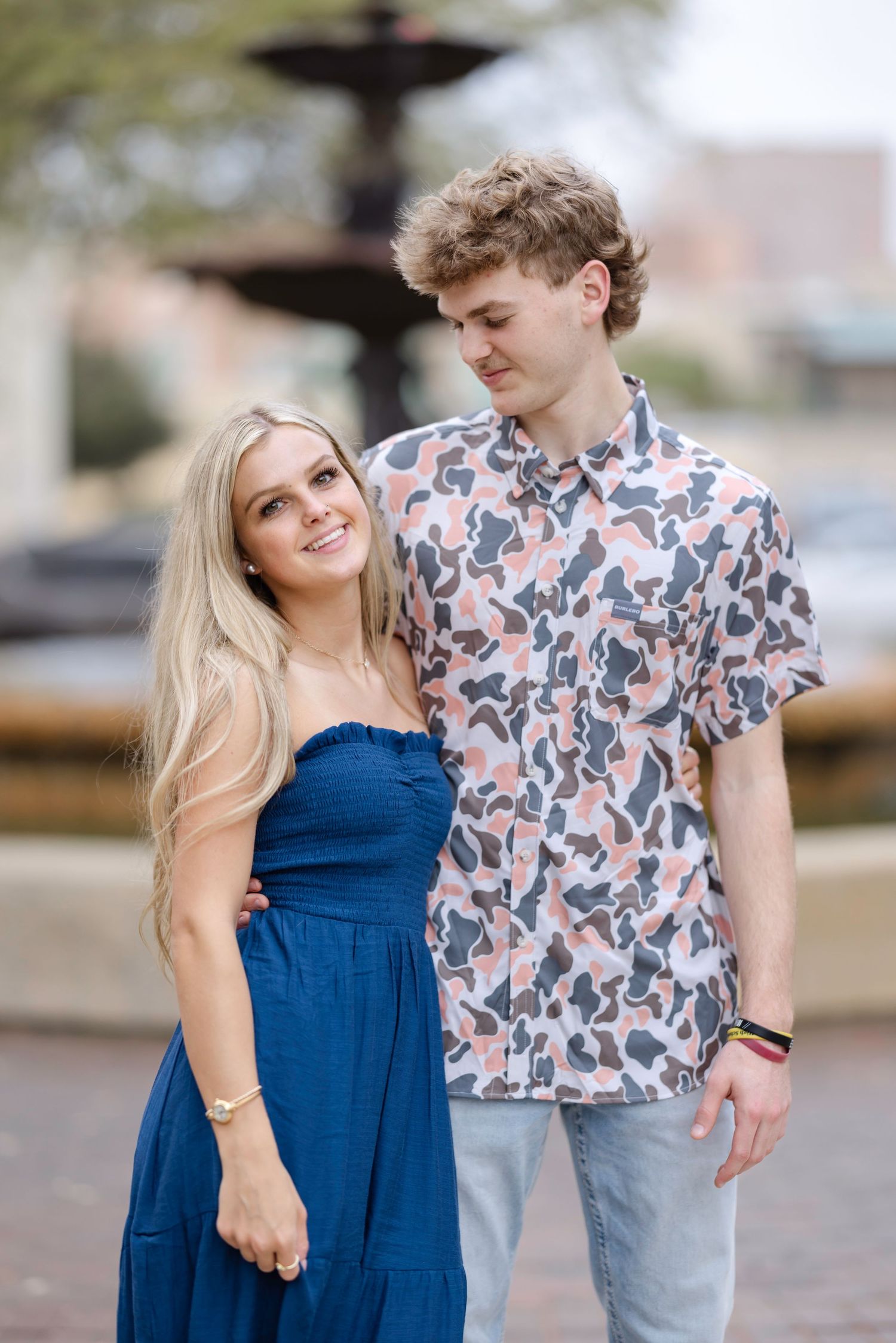 A couple poses together outdoors by a fountain, with her in a blue dress and him wearing a patterned short-sleeve shirt.