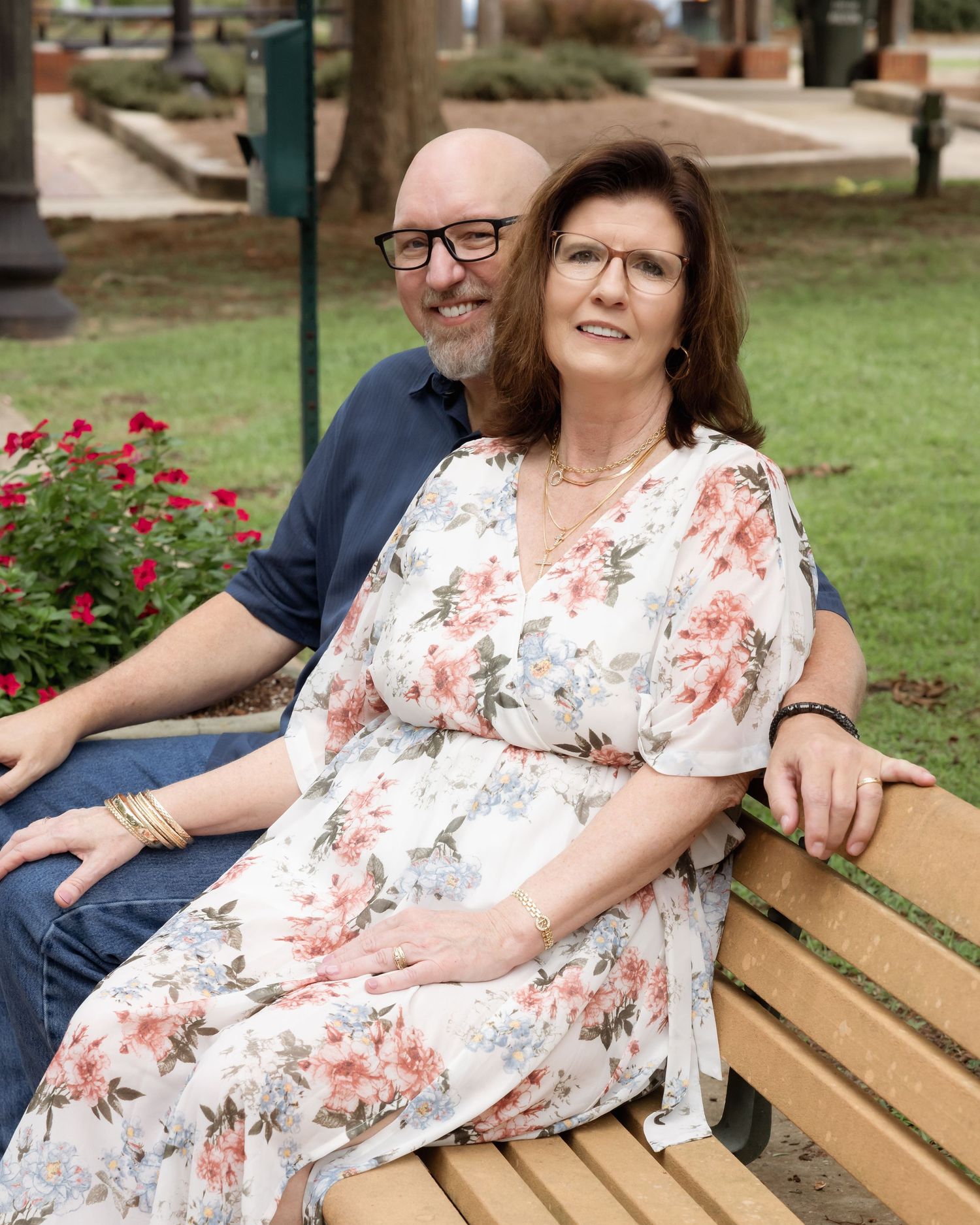 Couple sitting on a park bench surrounded by red flowers, woman in floral dress smiling warmly.