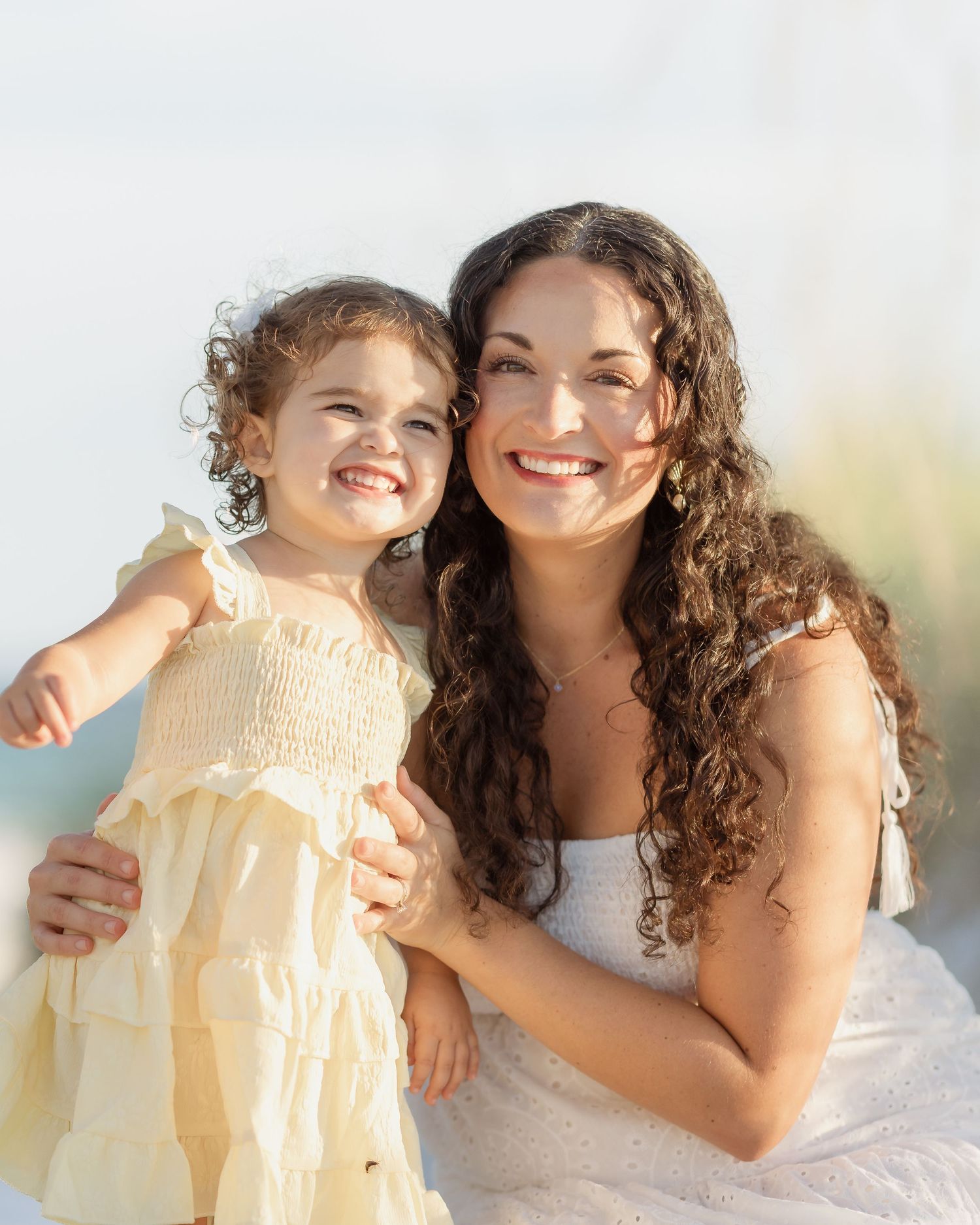A smiling woman with curly hair in a white dress holds a young girl in a yellow dress outdoors.