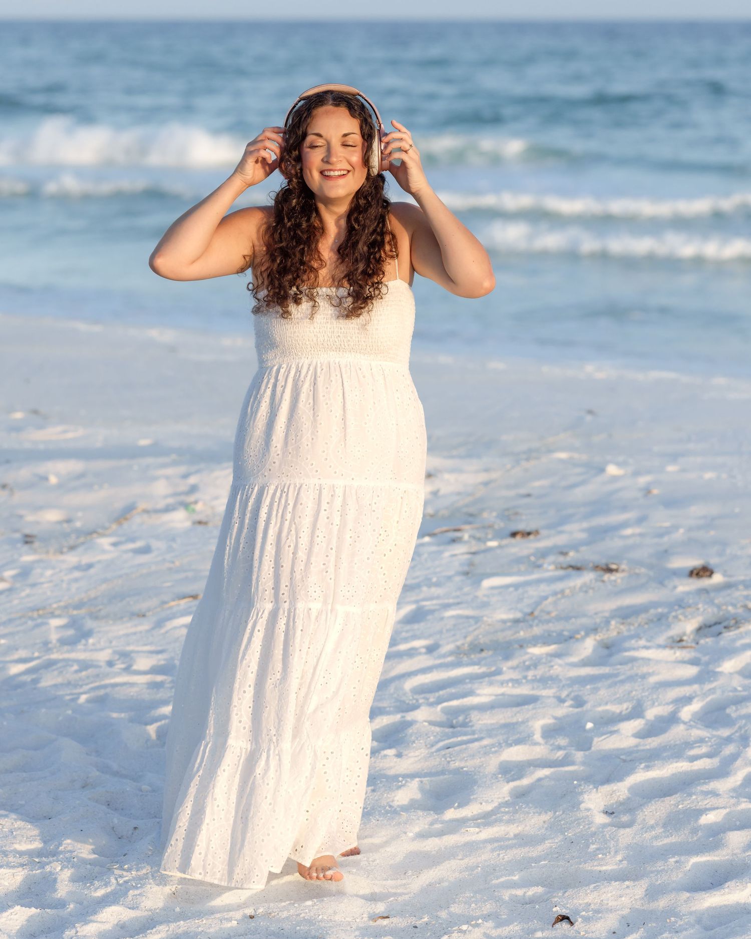 Happy woman in white dress adjusting headphones while standing on bright white sand beach.