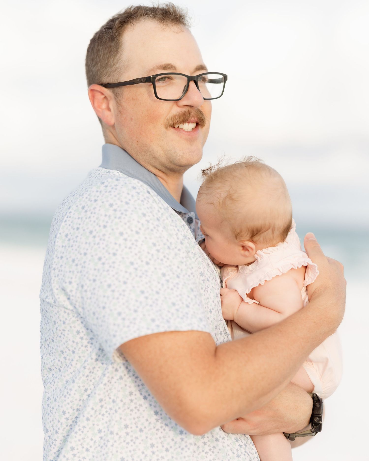 A man with glasses and a mustache holds a sleeping baby in a pink outfit on a beach.