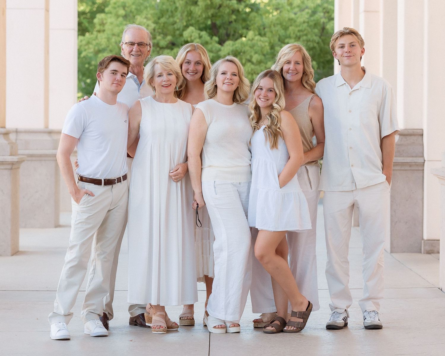 A happy family of seven posing together in coordinating white and beige outfits outdoors near elegant columns.