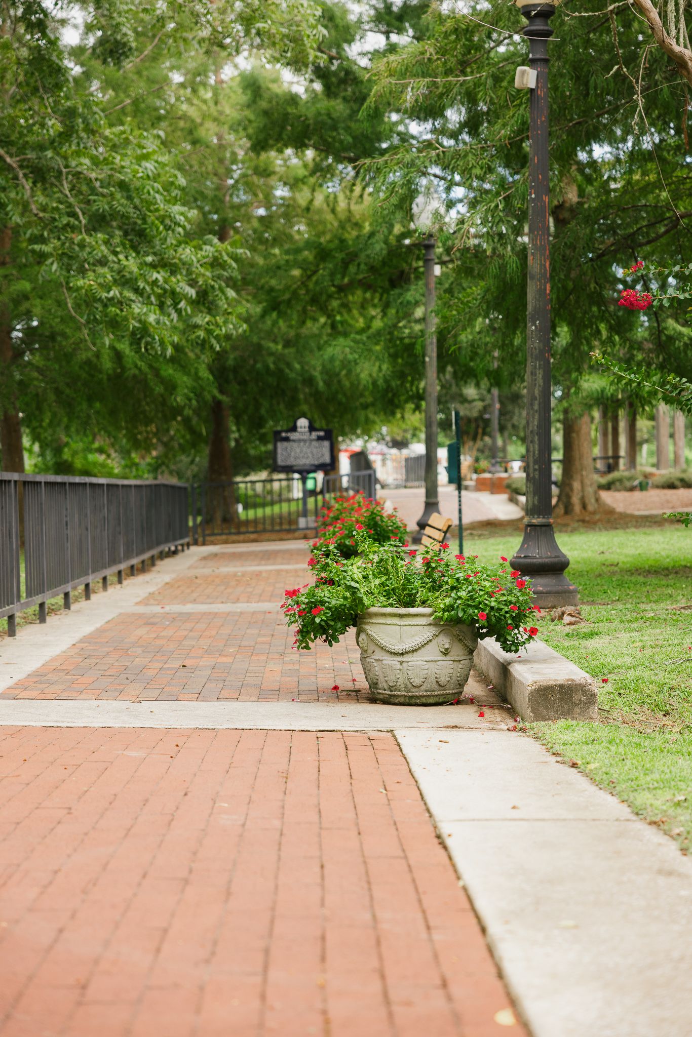 Brick pathway in a lush park with a large flower pot, lamp post, and green trees in the background.