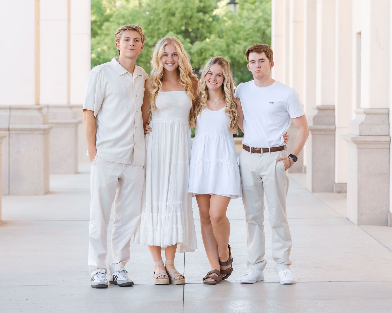 Four young adults in coordinated white outfits posing together between columns outdoors.