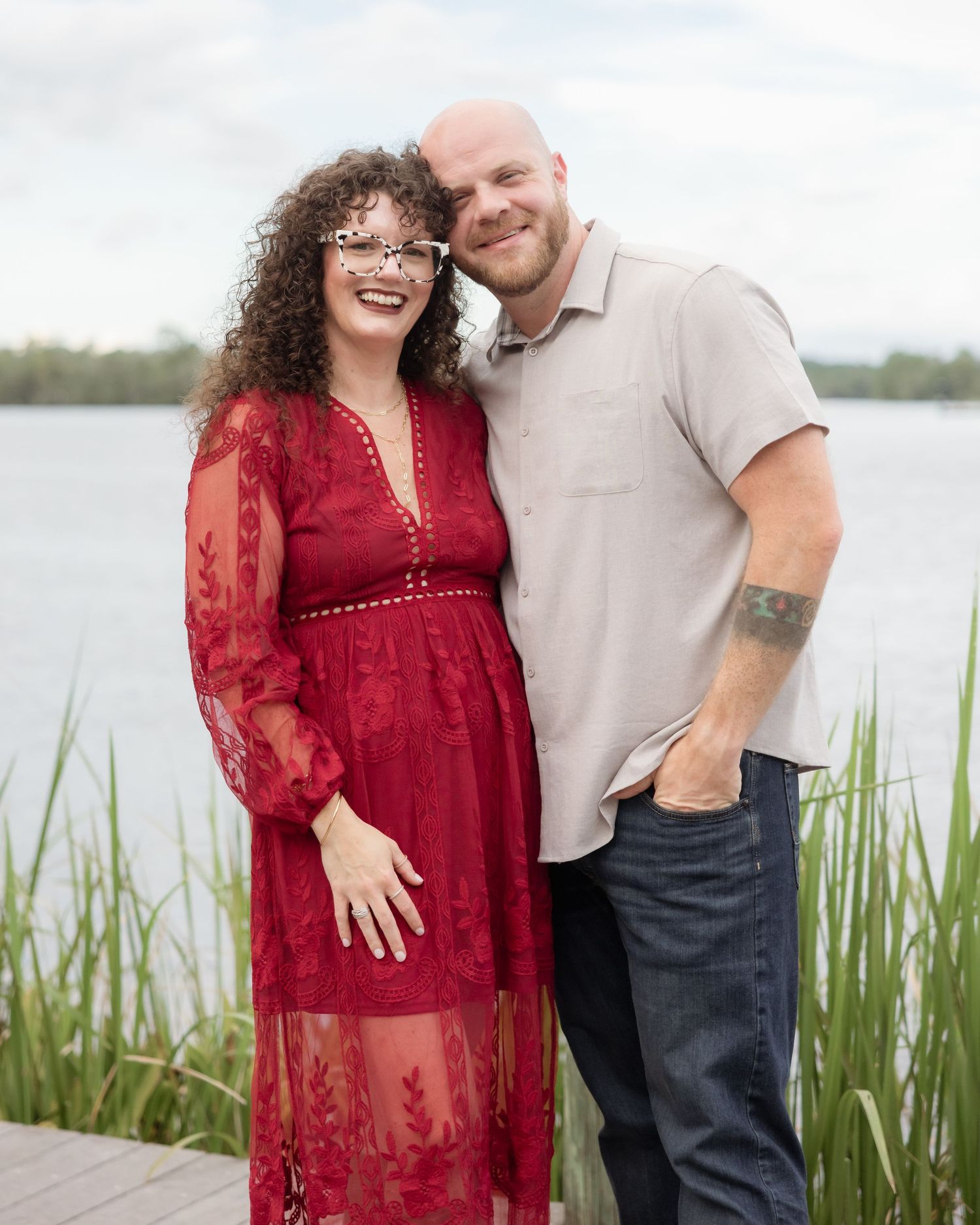 Couple smiling together on a lakeside dock, woman in red lace dress and man in gray shirt.