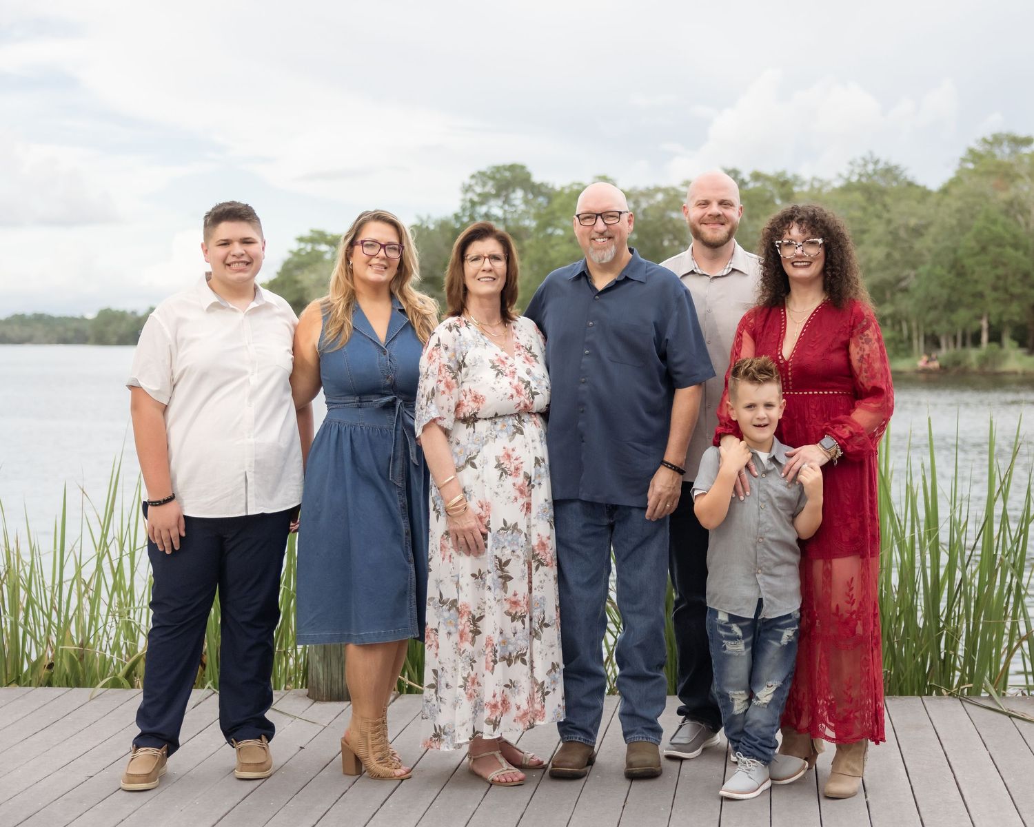 Family of seven posing together on a wooden dock by a lake, smiling outdoors on a cloudy day.