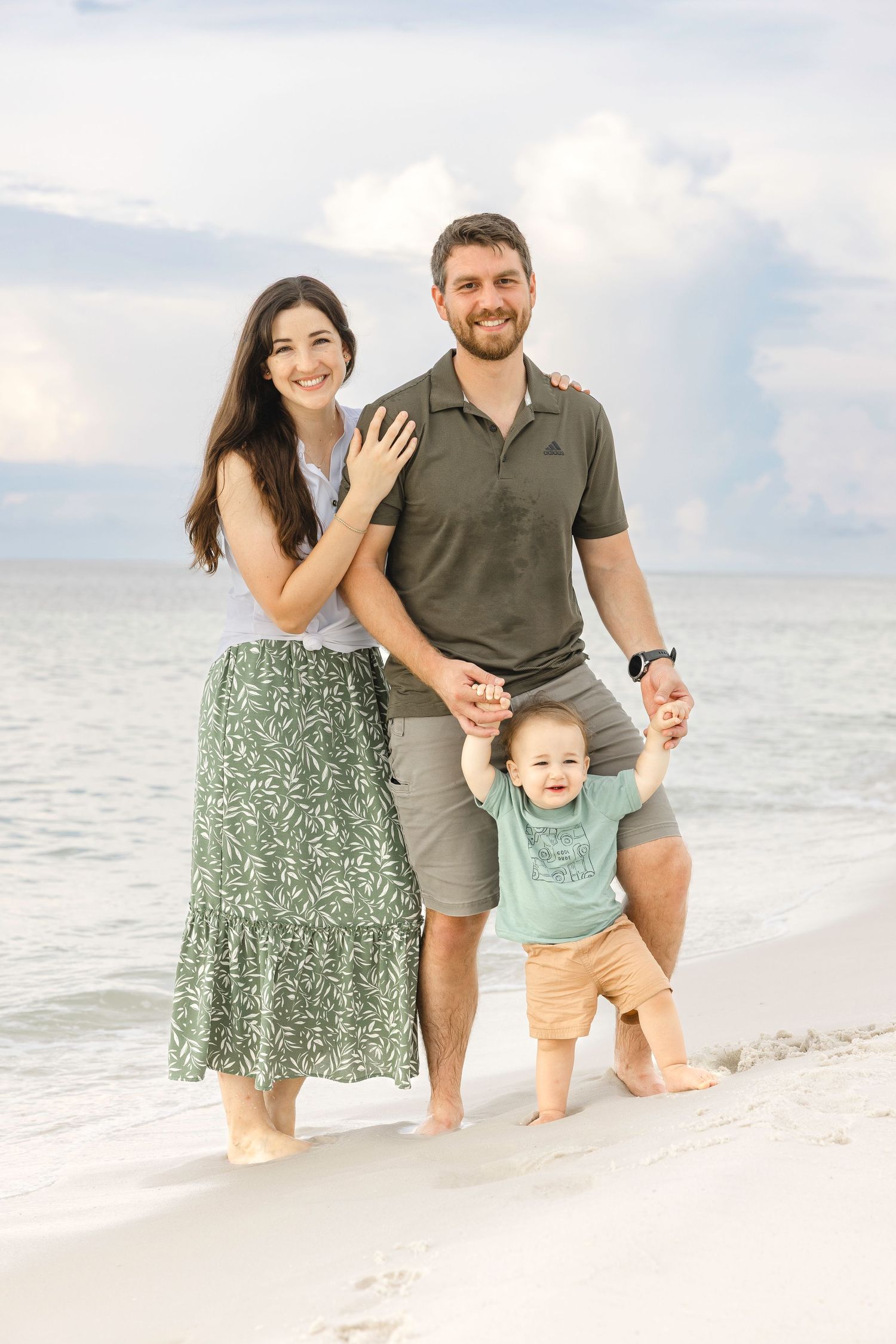 A family stands together smiling on a sunny beach with calm ocean waters and white sand in the background.