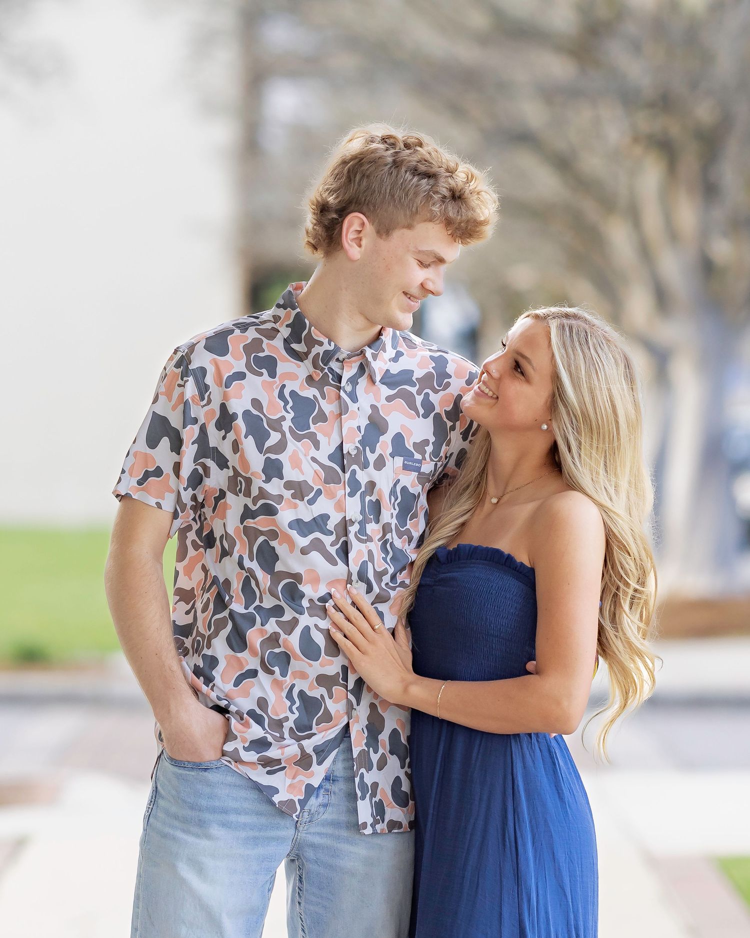 A couple in casual attire sharing a romantic moment on a path lined with trees in a park setting.