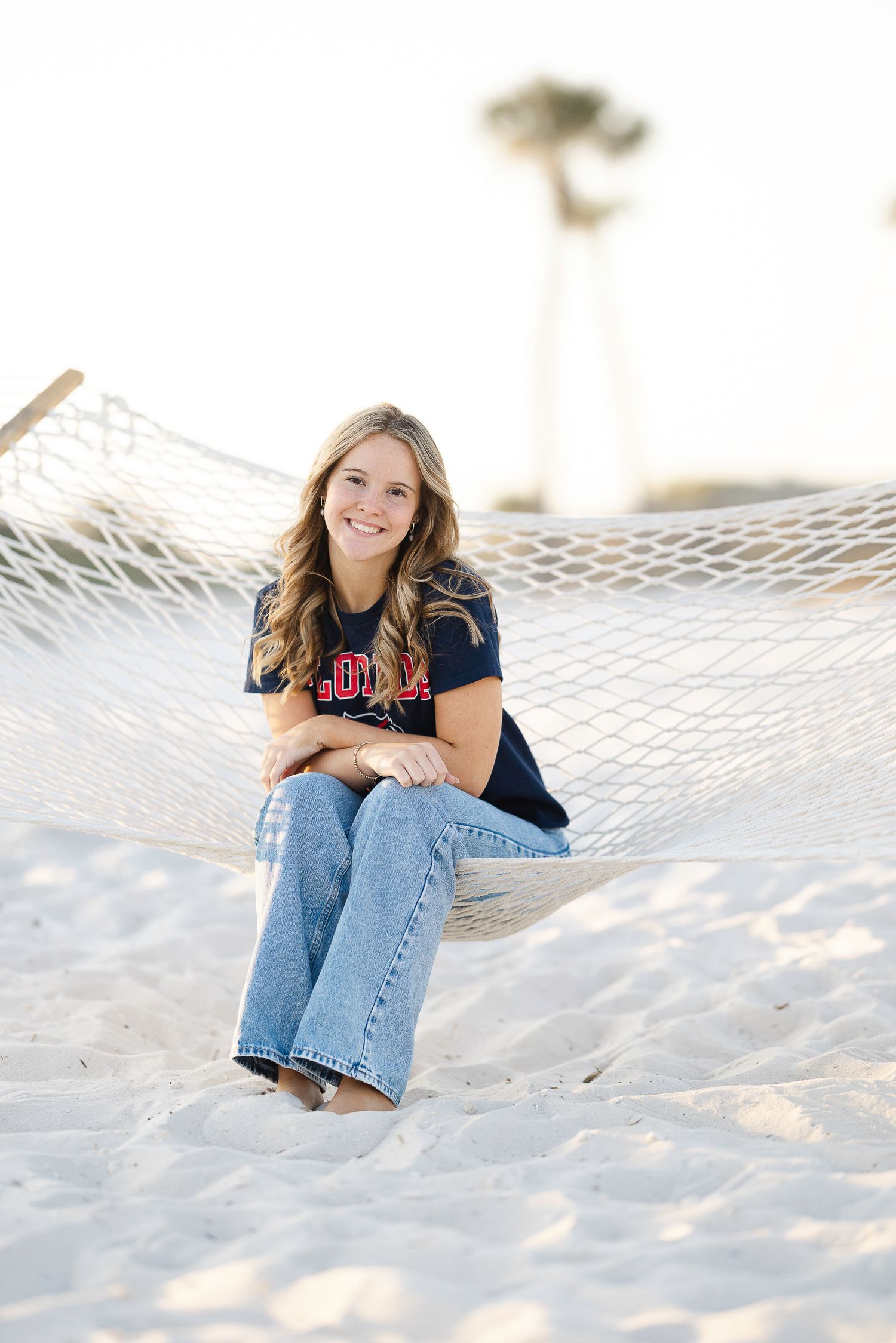 A person in a navy blue shirt and jeans sits casually on pristine white sand at the beach.