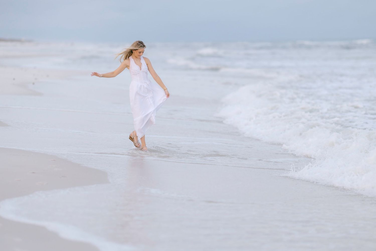 A person in a flowing white dress runs gracefully along the beach shoreline as waves roll in during golden hour.