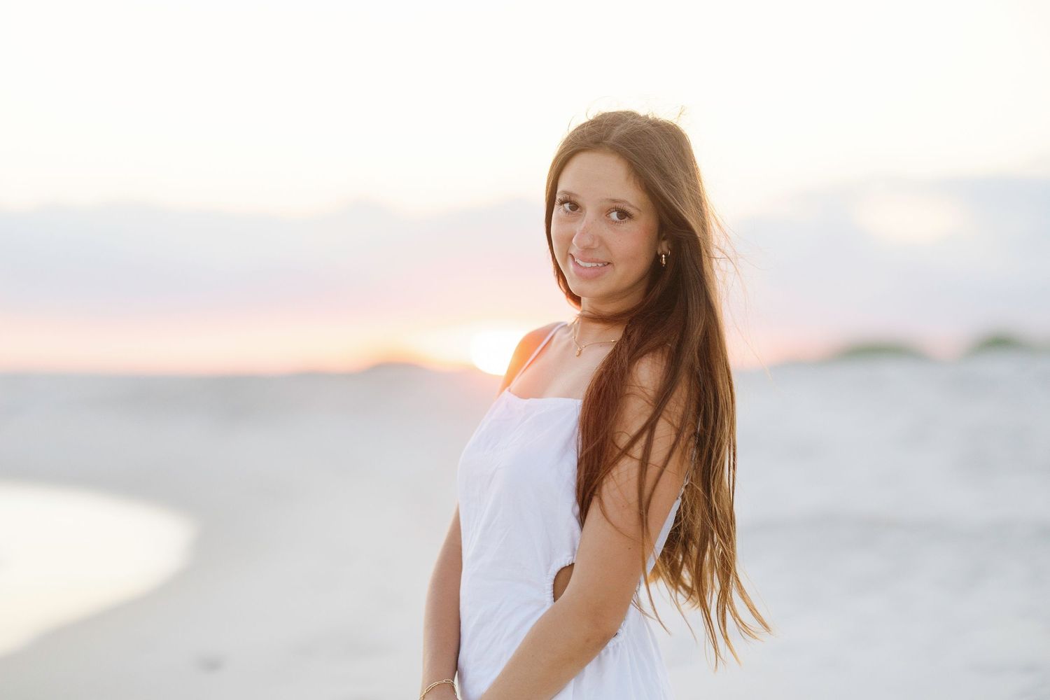 A person in white beachwear poses at sunset with the ocean and sand dunes creating a scenic backdrop.