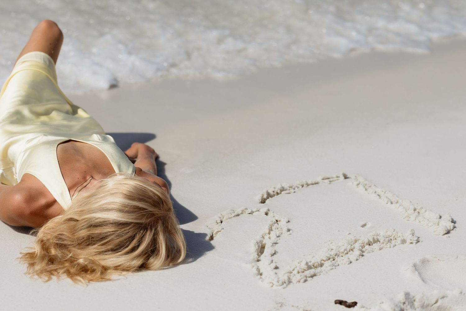 Blonde woman lying on sandy beach shore with heart drawn in sand.