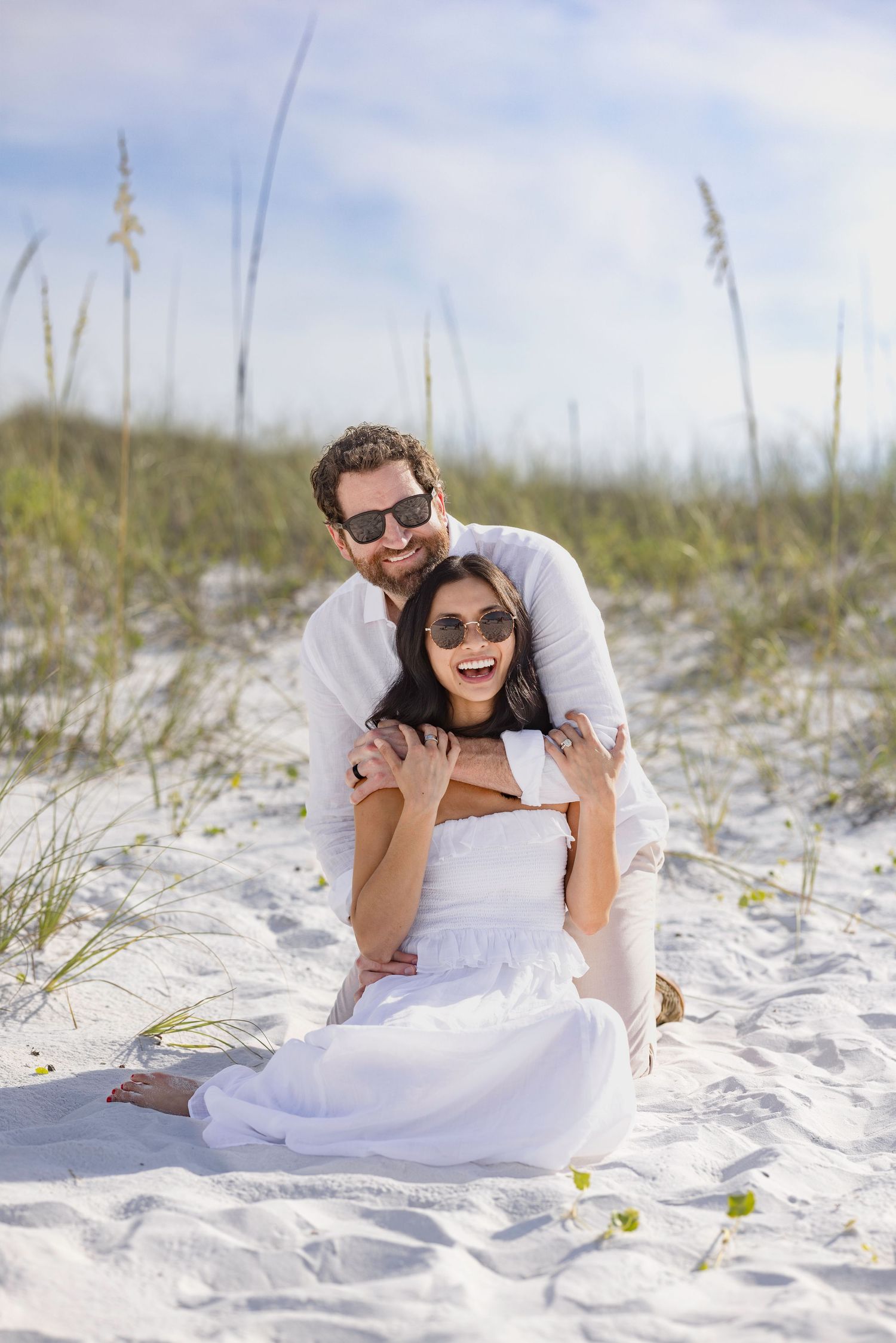 Happy couple in white outfits sitting on sandy beach dunes, smiling and wearing sunglasses on a sunny day.