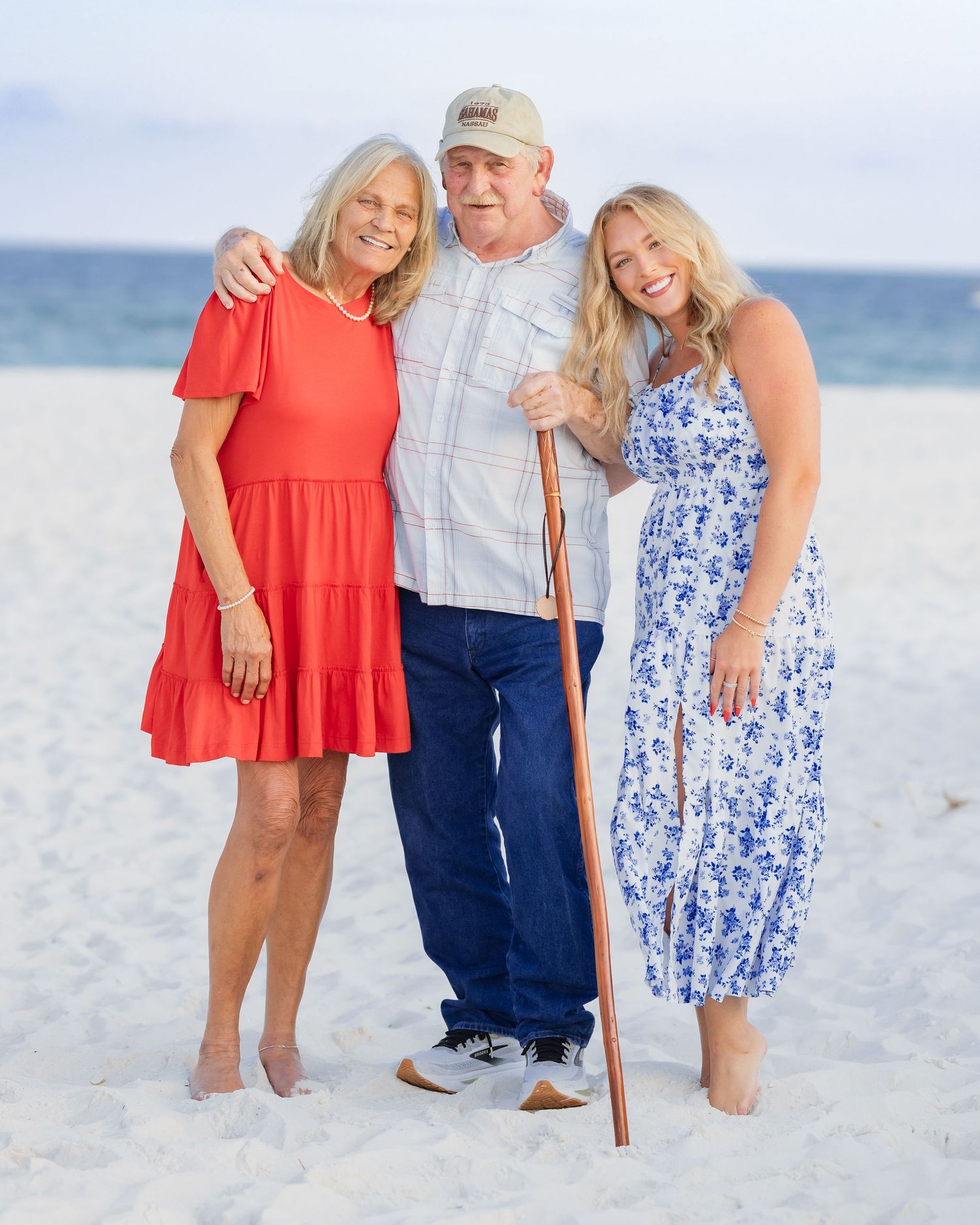 Three people posing together on a sandy beach, man with cane flanked by two smiling women in summer dresses.