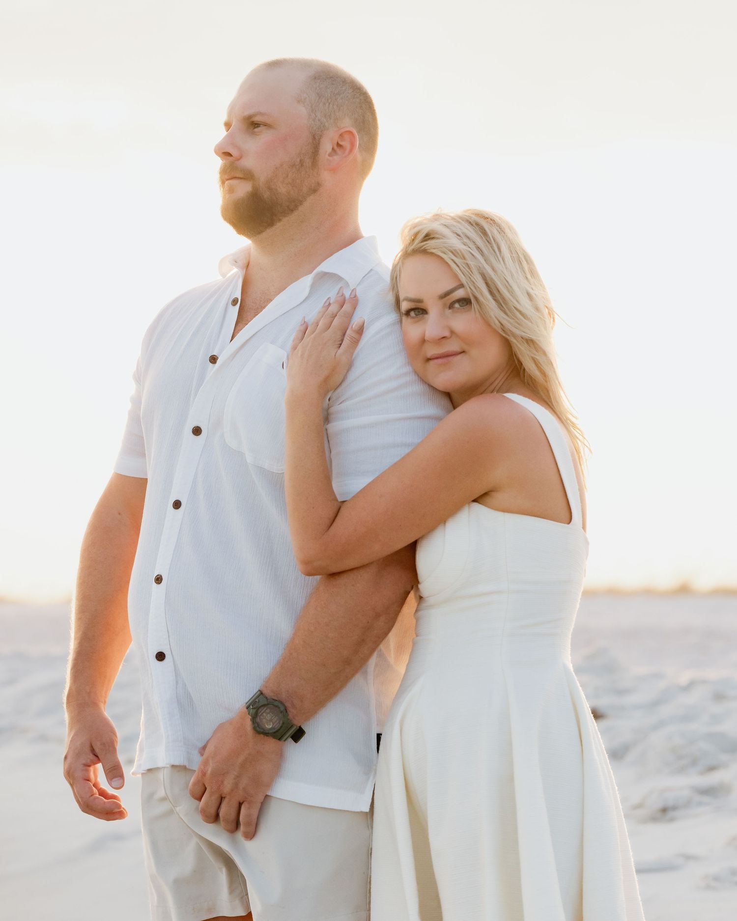 Romantic couple in white outfits posing back to back on a sunlit beach at golden hour.