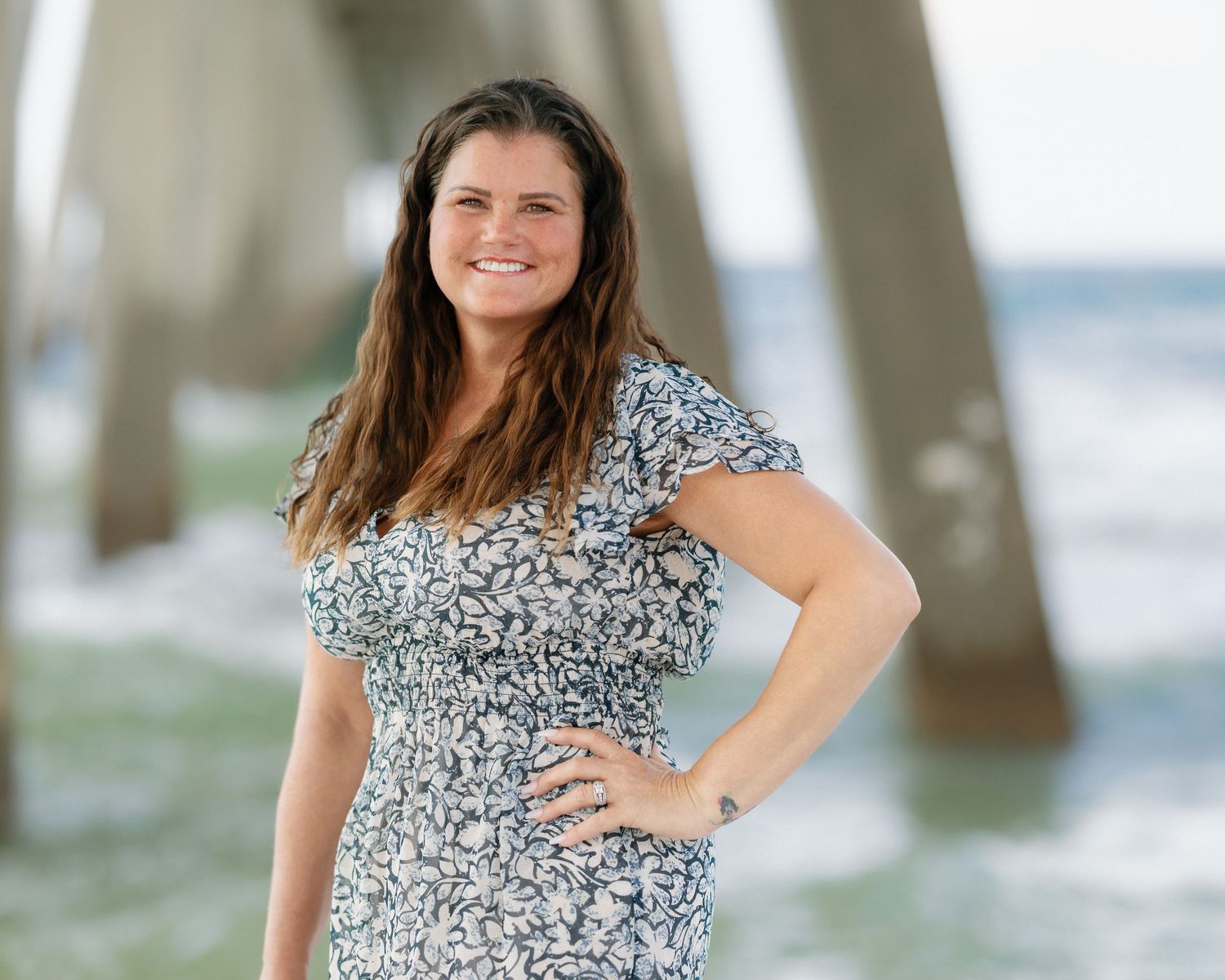 Smiling woman in floral dress posing confidently on beach near pier on a bright sunny day.