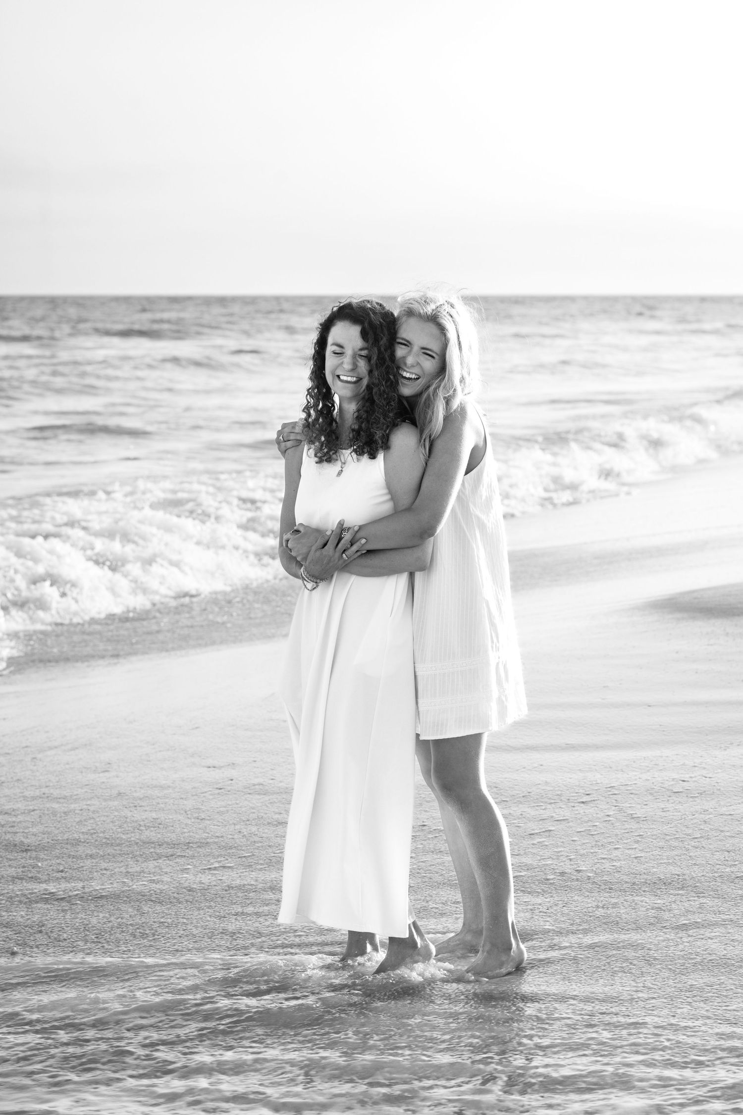 Two women in white dresses laugh and embrace on a beach shoreline in a black and white photo.
