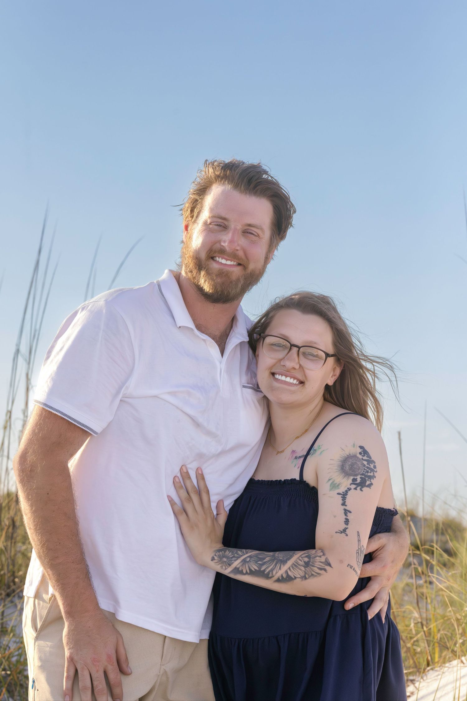 Happy couple embracing on beach dunes, woman with tattoos and glasses in navy dress, man in white polo shirt, clear blue sky.