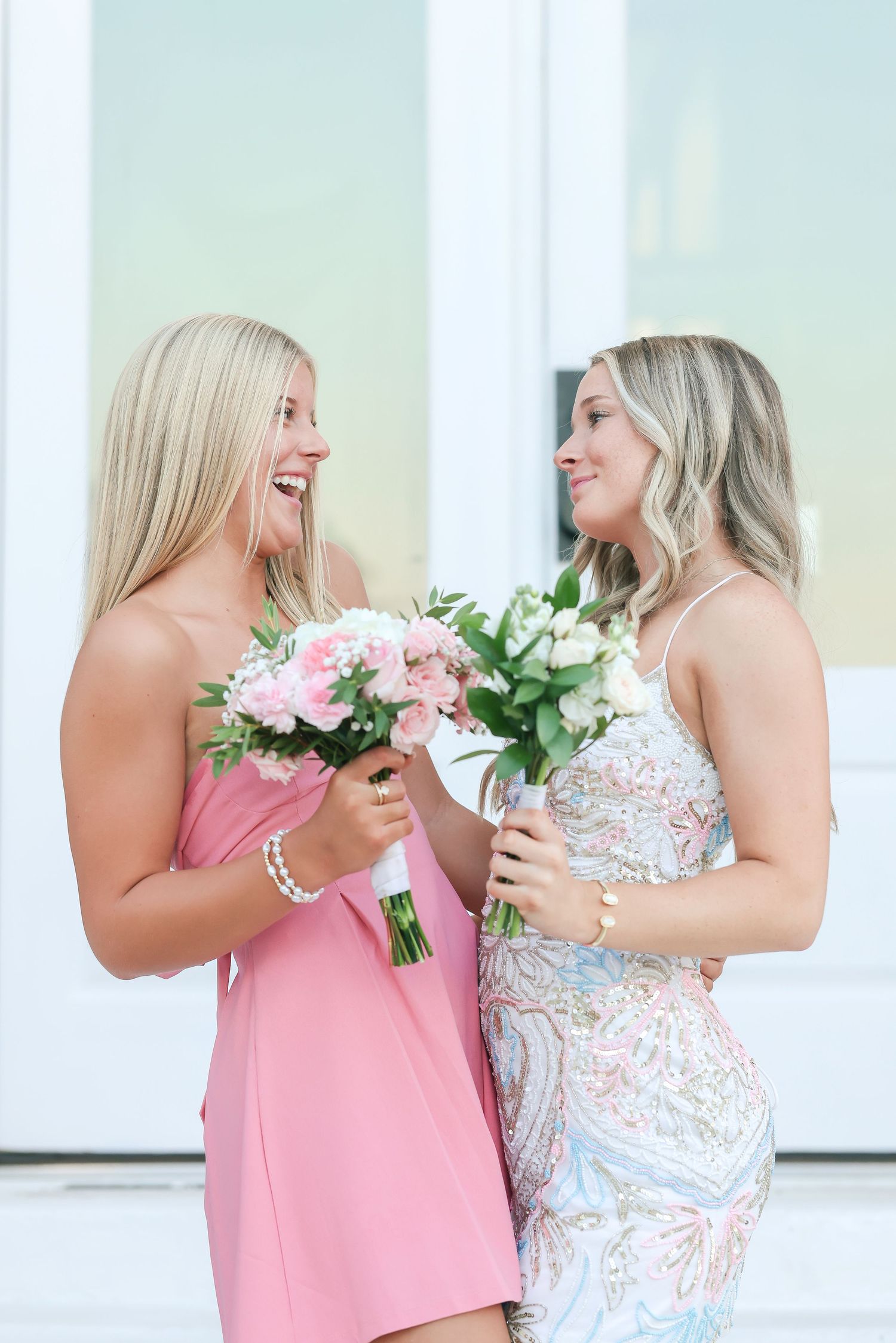 Two smiling blonde women in formal dresses holding pink and white flower bouquets at a special occasion.