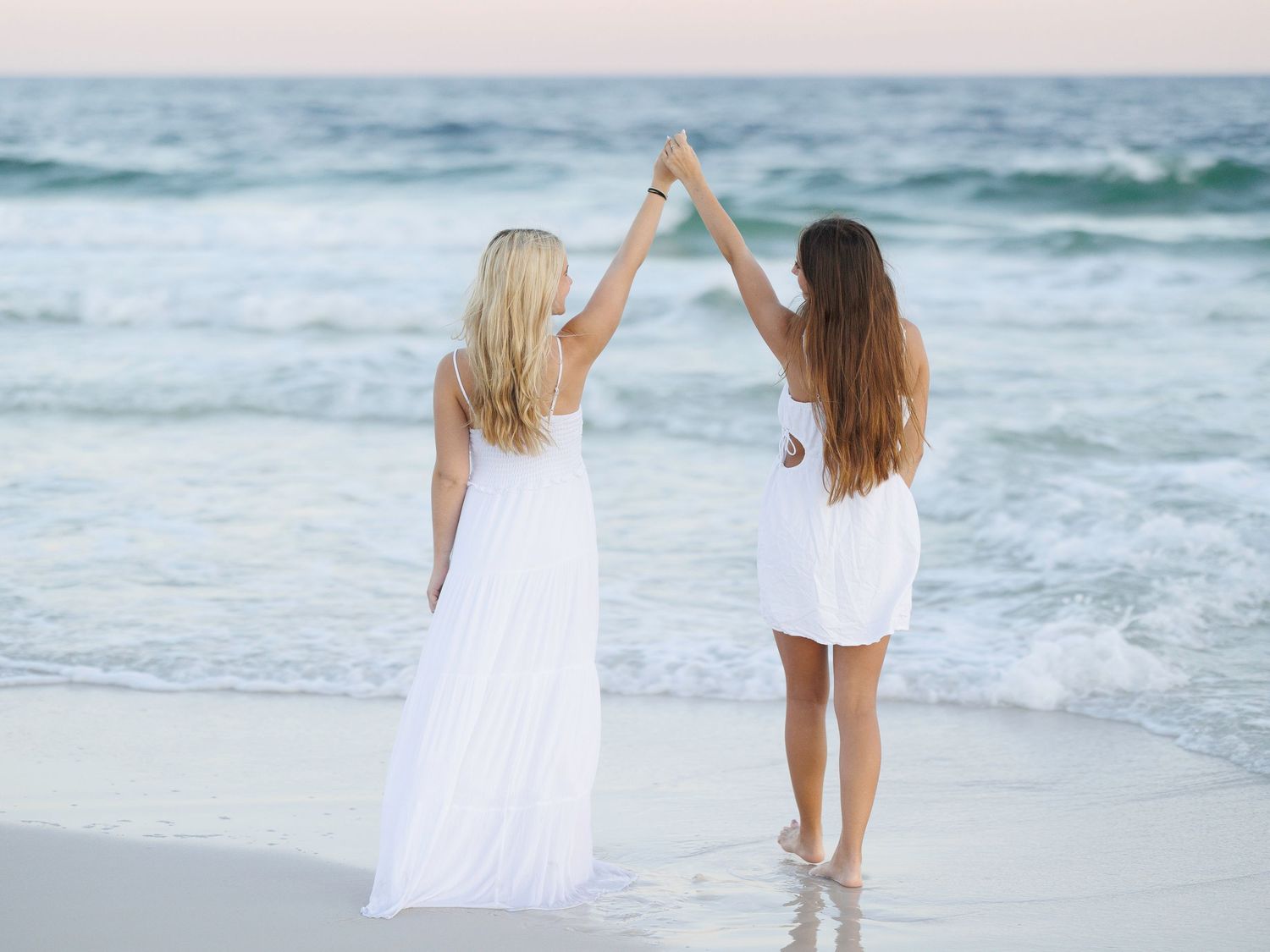 Women in white dresses dancing and playing at the beach edge.