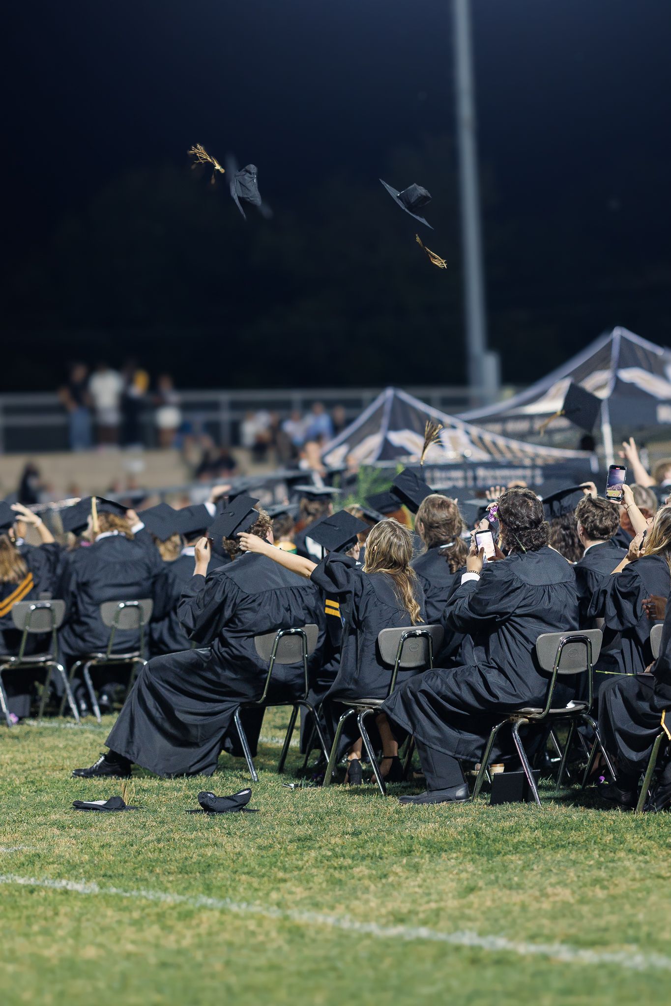 Graduation caps fly through the night sky as graduates celebrate during outdoor ceremony.
