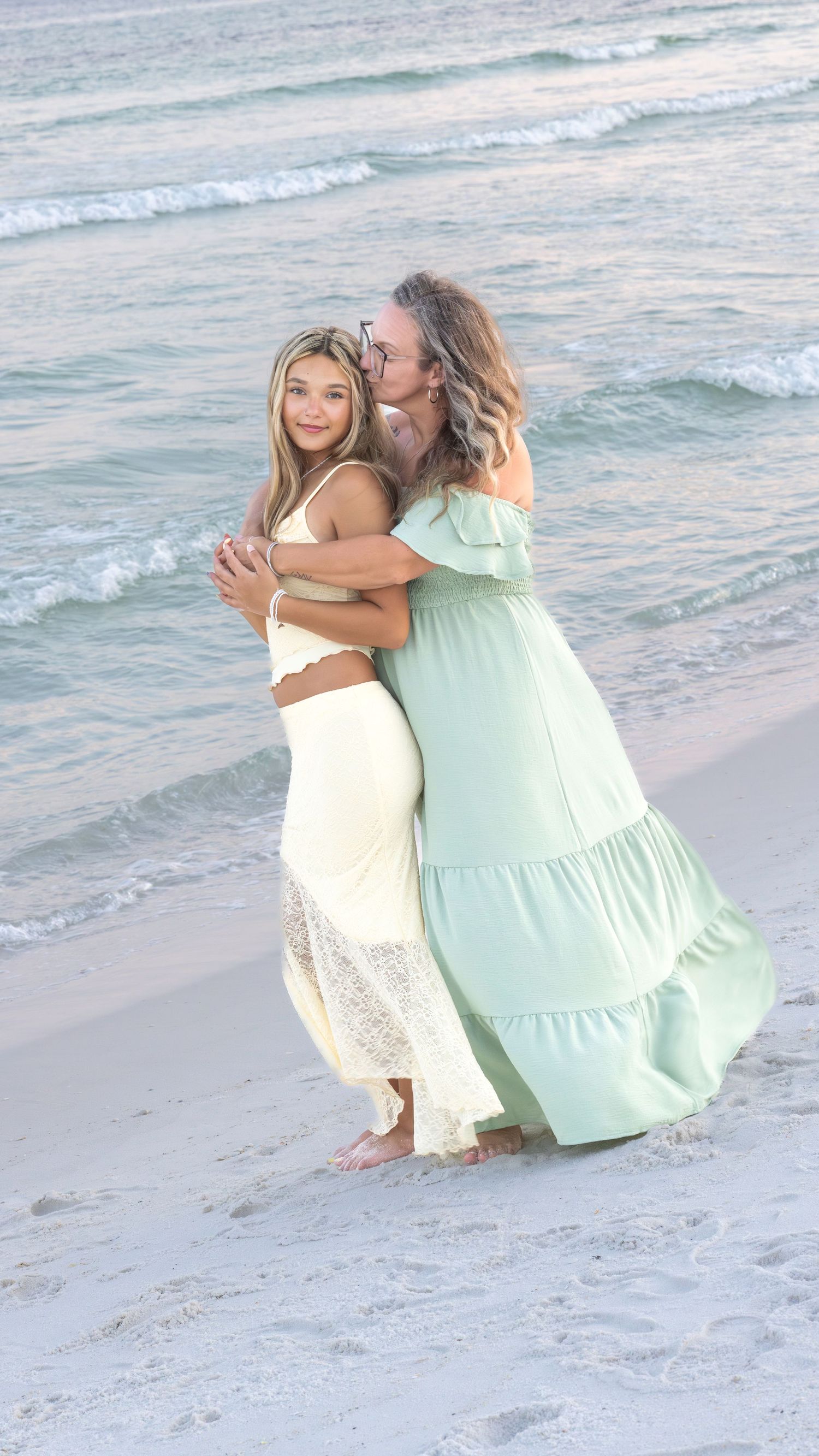 Two women embrace on a sandy beach at sunset, wearing flowing white and mint green dresses with ocean waves behind them.