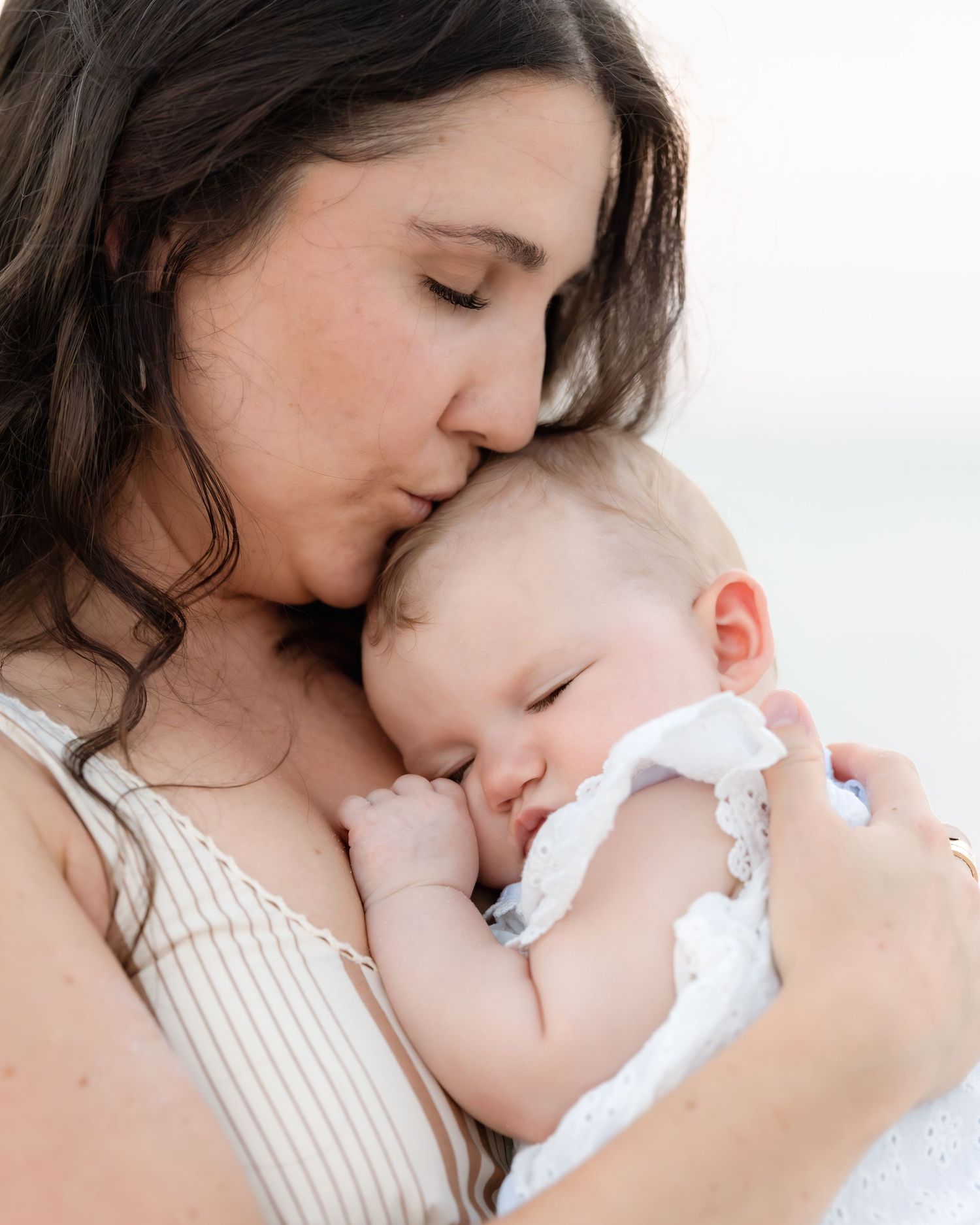 Loving mother with dark hair gently kissing sleeping newborn baby at the beach.