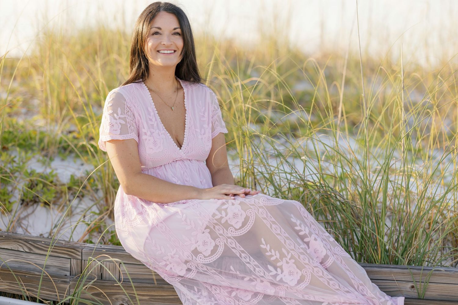 A person in a pink lace dress sits on sandy dunes surrounded by beach grass during golden hour.