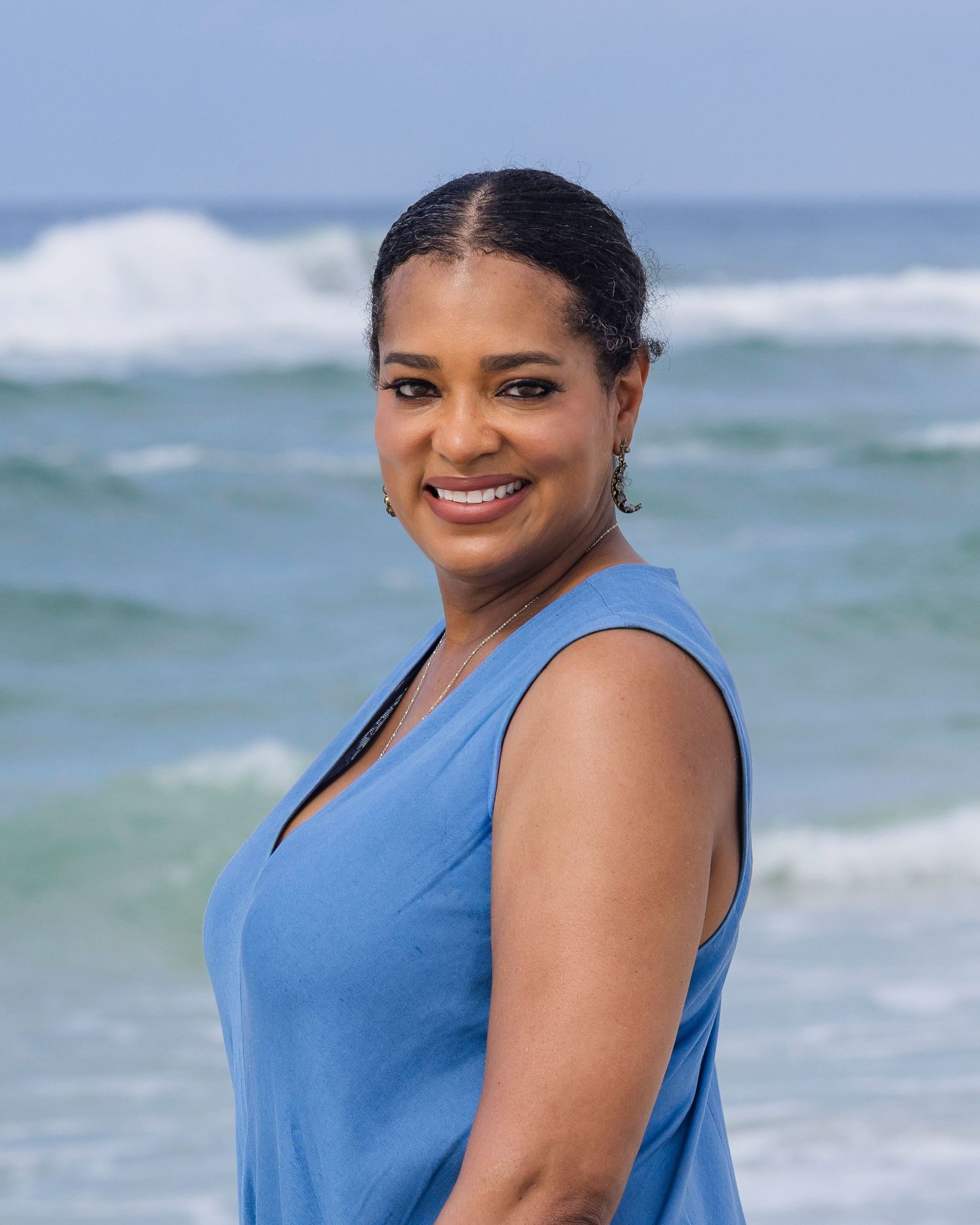 Smiling woman in blue sleeveless top posing on a beach with turquoise ocean waves in the background.