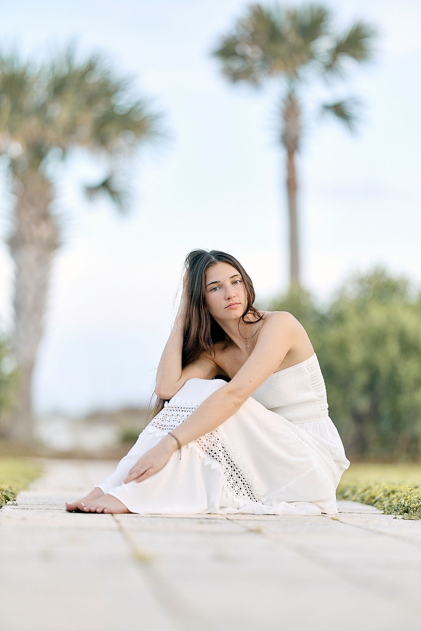 Young woman in white dress sitting on wooden boardwalk with palm trees in background.