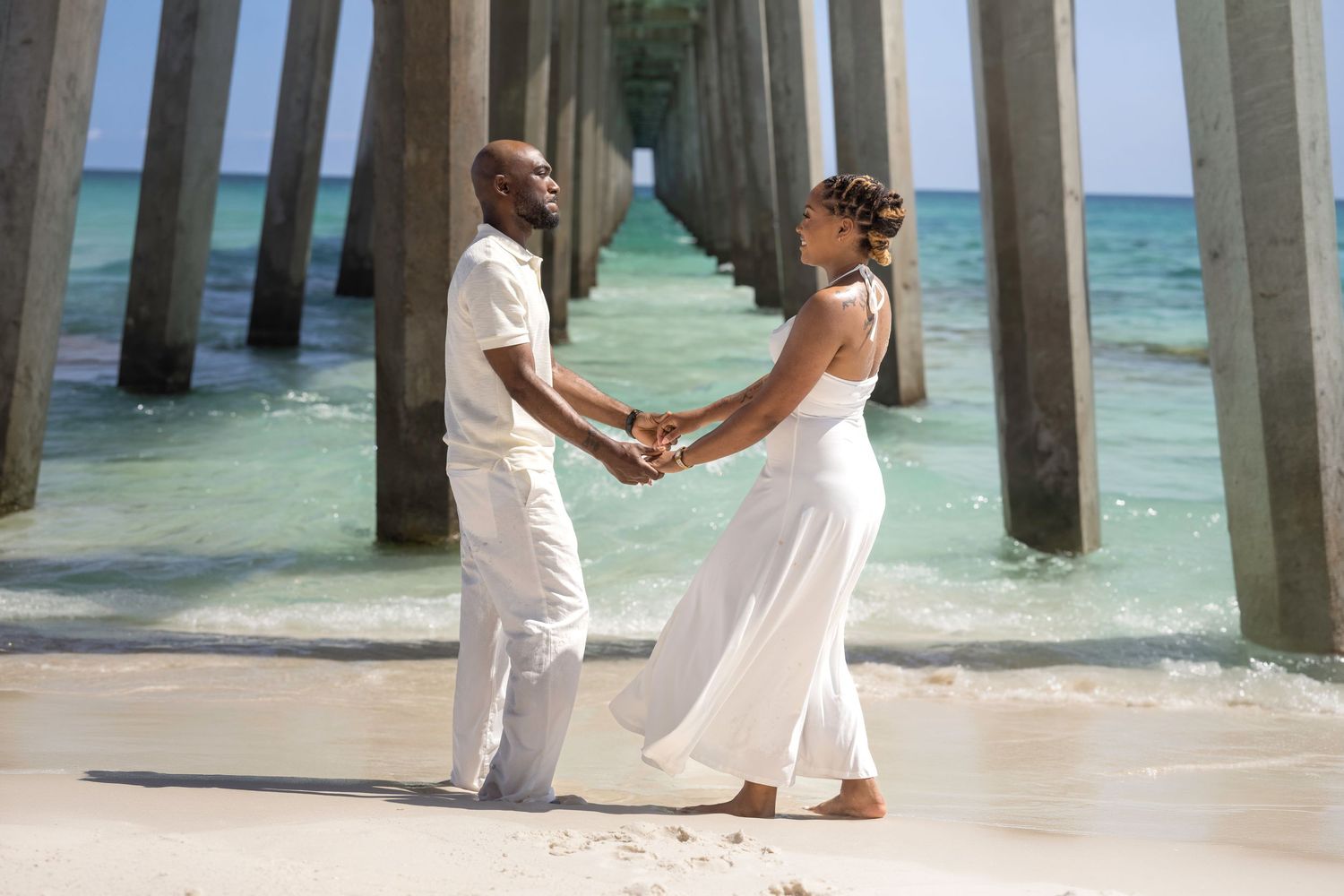 Couple in white attire holding hands on a beach under a pier with turquoise water in the background.