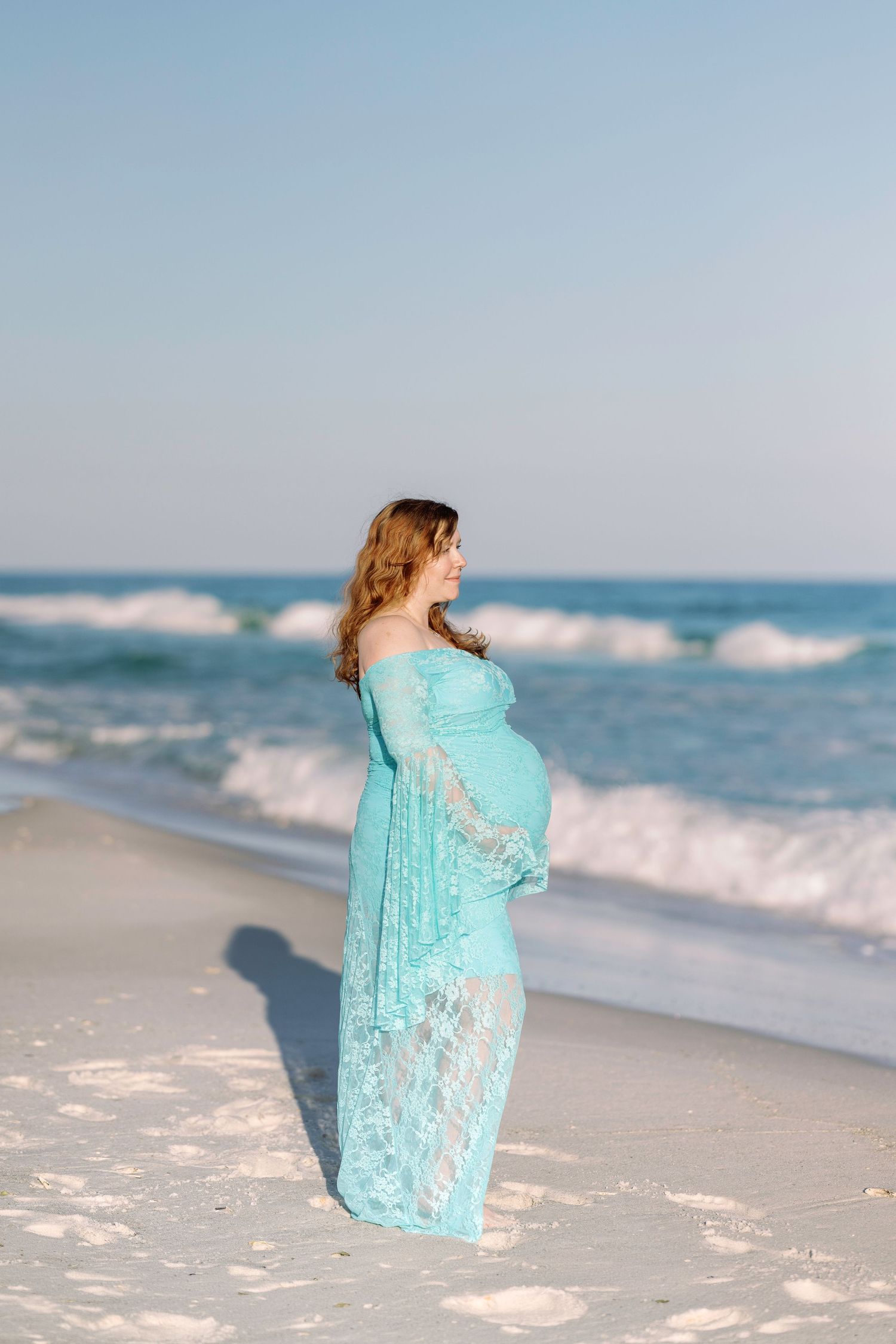 Pregnant woman in teal lace off-shoulder gown standing on white sand beach with turquoise ocean waves behind her.