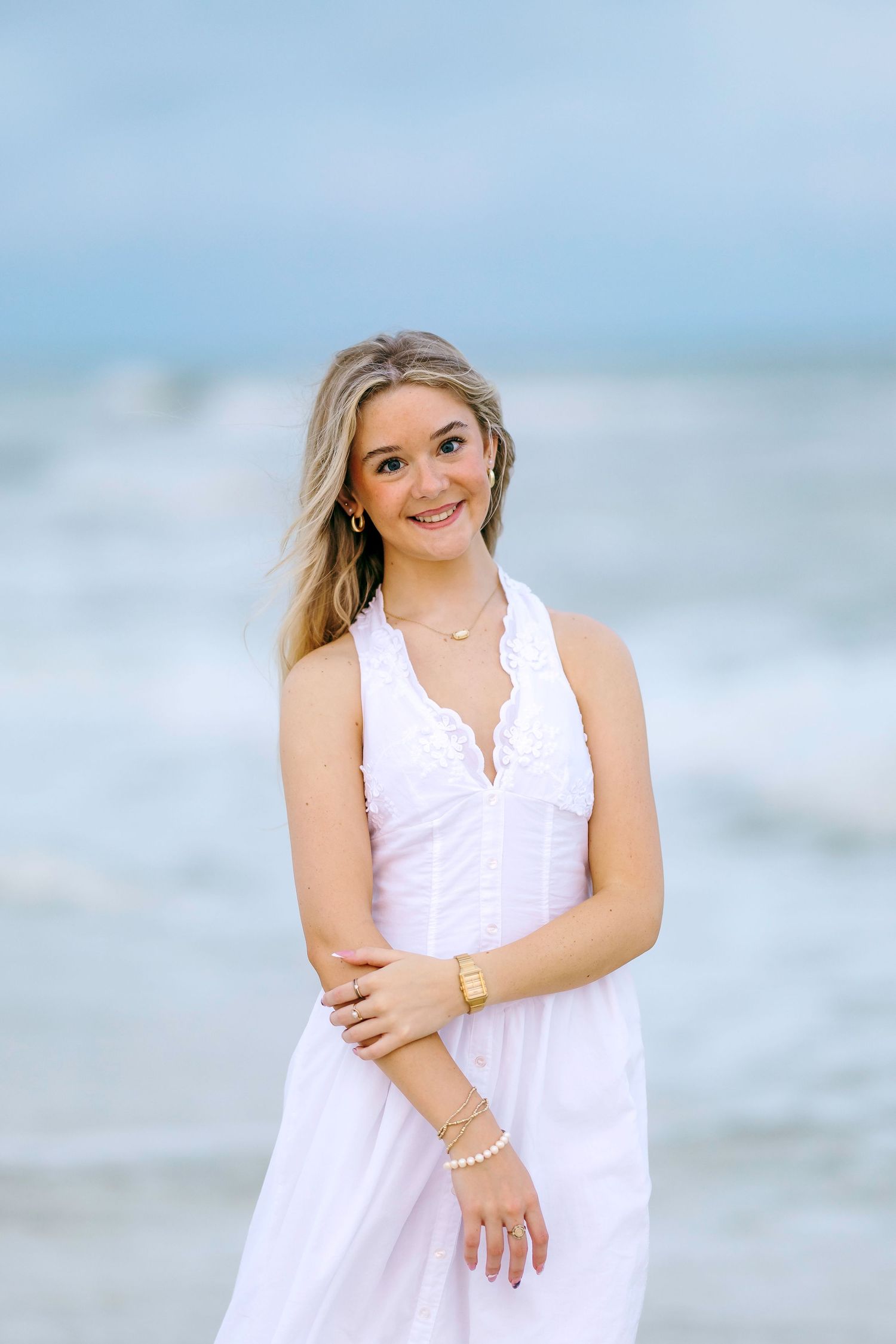 Young blonde woman in white halter dress smiling at beach with ocean waves behind her.