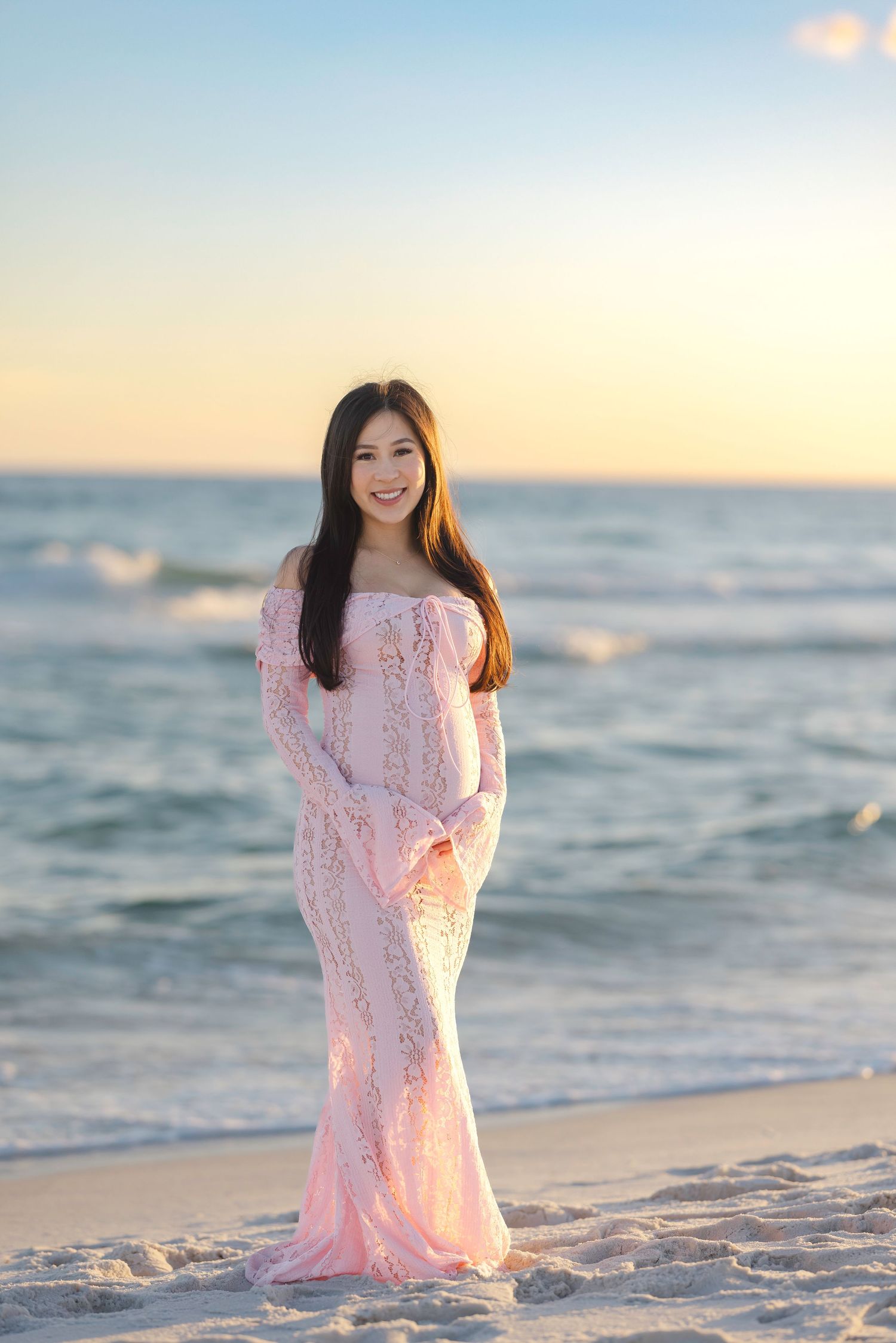 A young woman in a pink lace off-shoulder gown poses gracefully on a sandy beach at sunset.